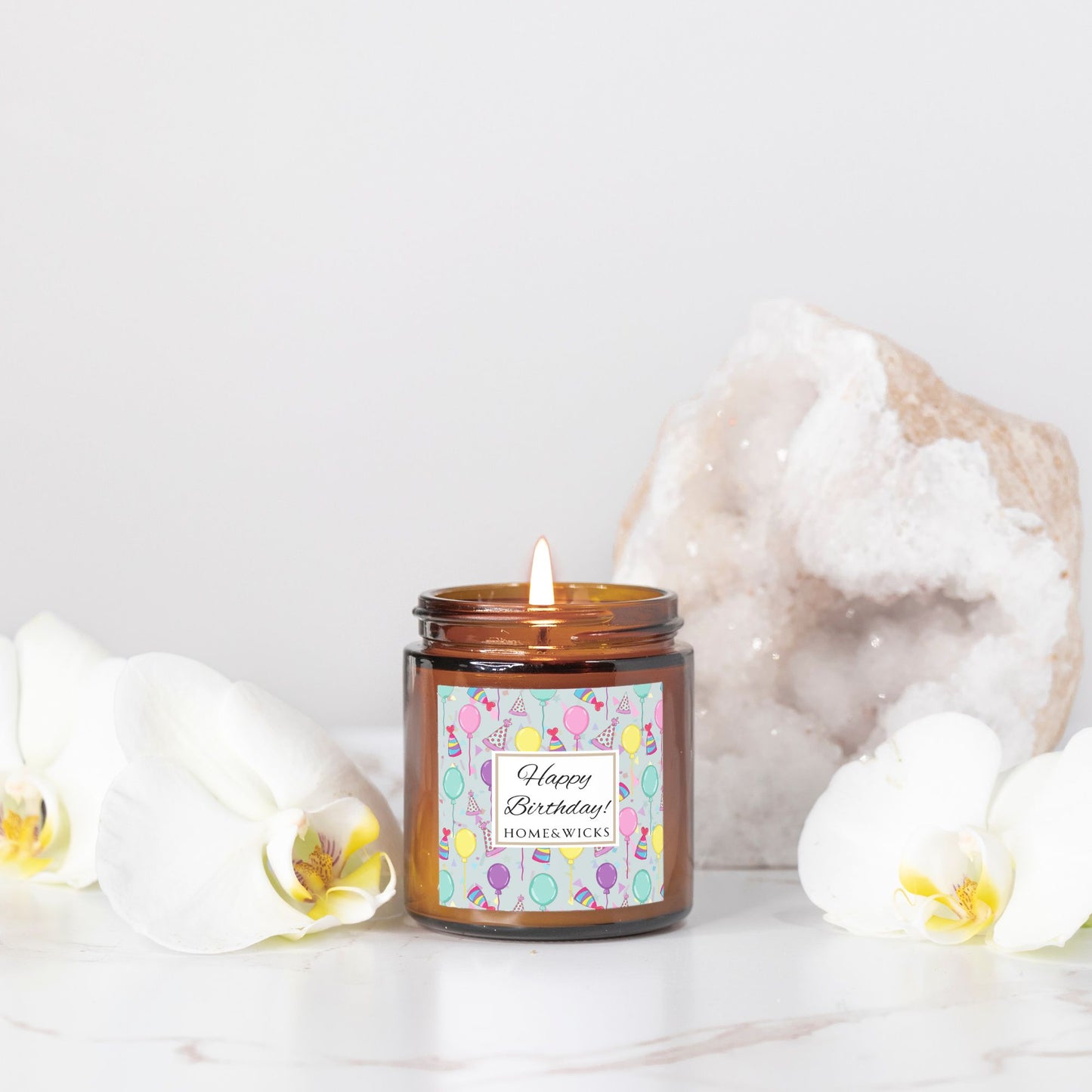 Amber candle jar with gold lid with a 'Happy Birthday' label featuring balloons, birthday party hats and confetti on a light background.