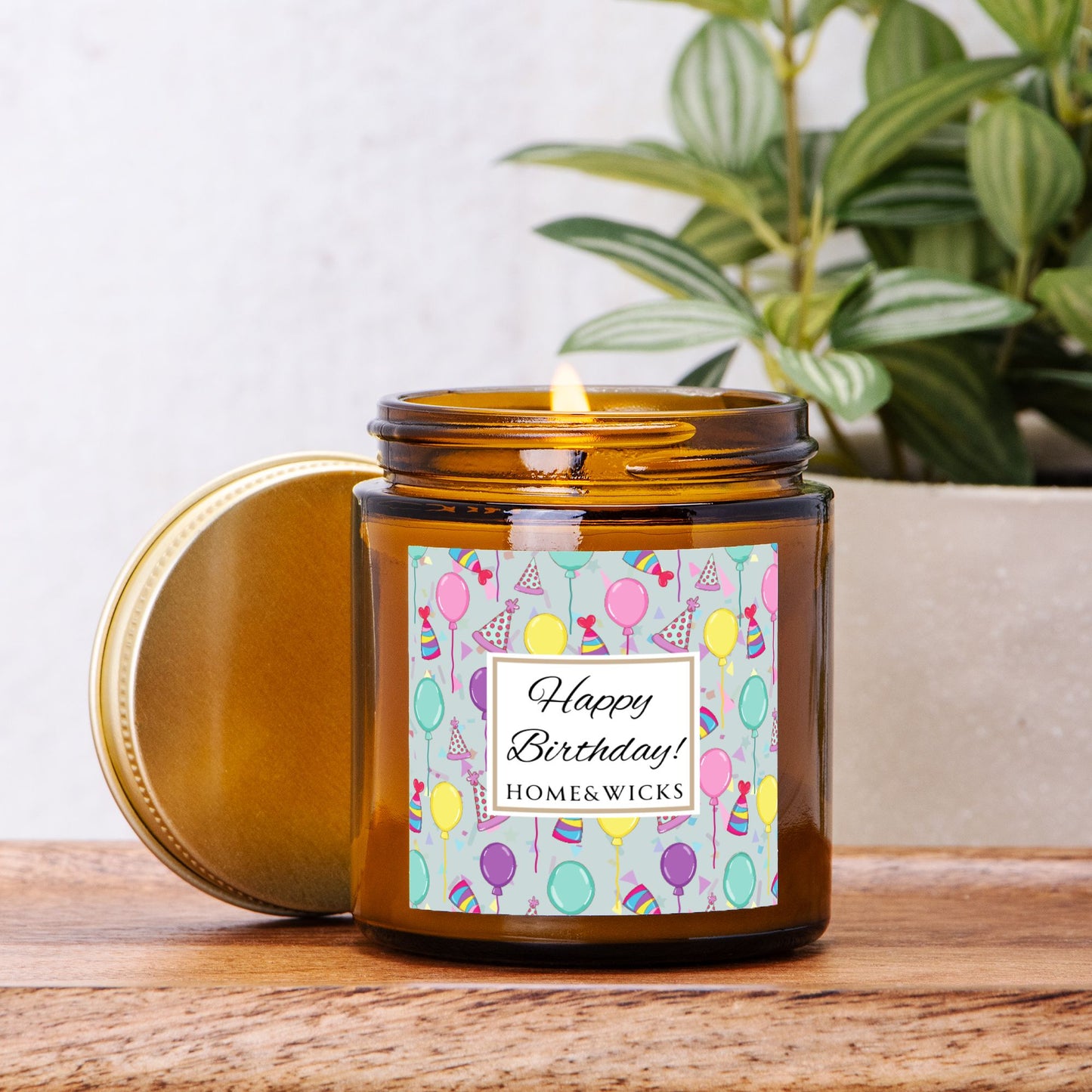 Amber candle jar with gold lid with a 'Happy Birthday' label featuring balloons, birthday party hats and confetti on a wooden surface with a plant in the background