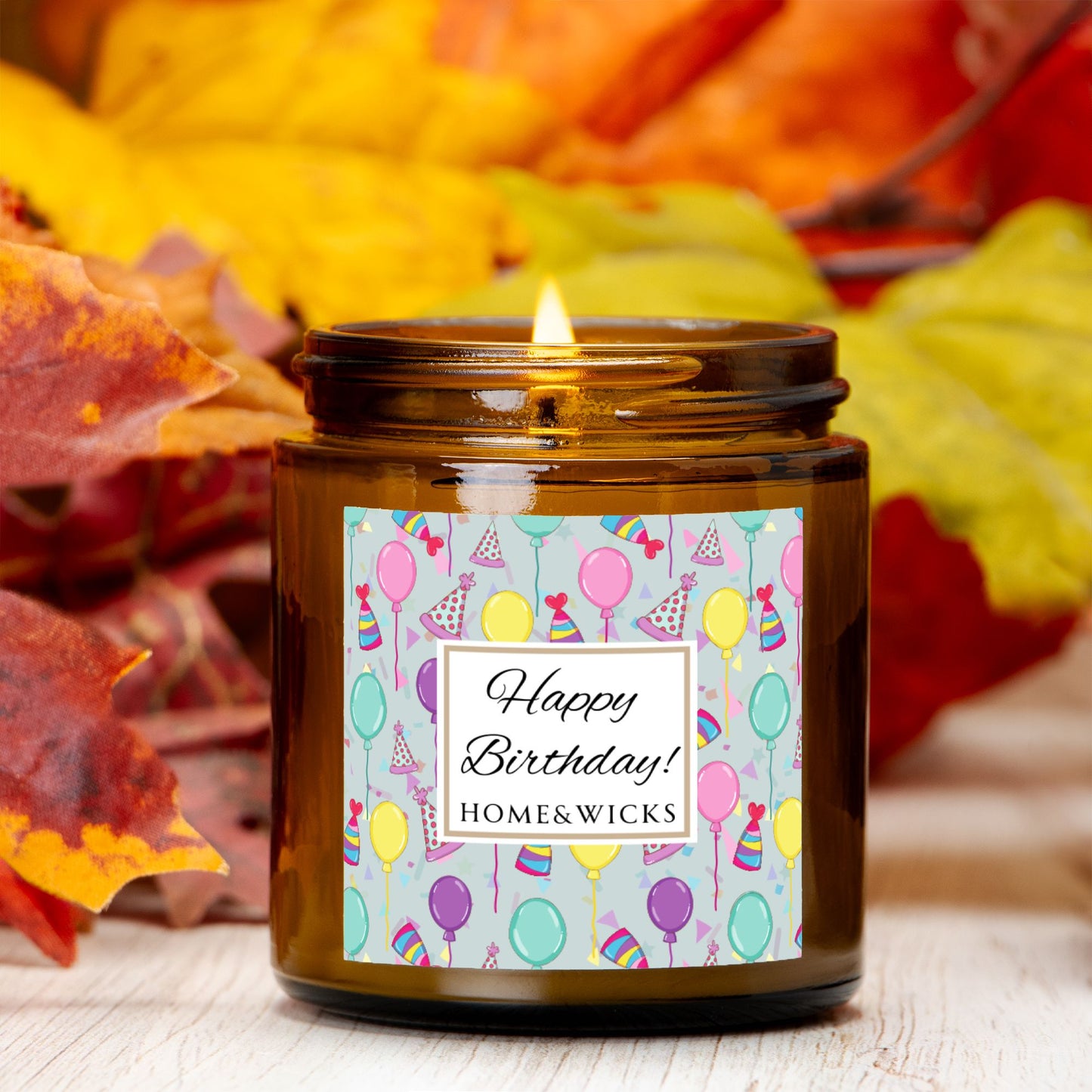 Amber candle jar with gold lid with a 'Happy Birthday' label featuring balloons, birthday party hats and confetti surrounded by autumn leaves.