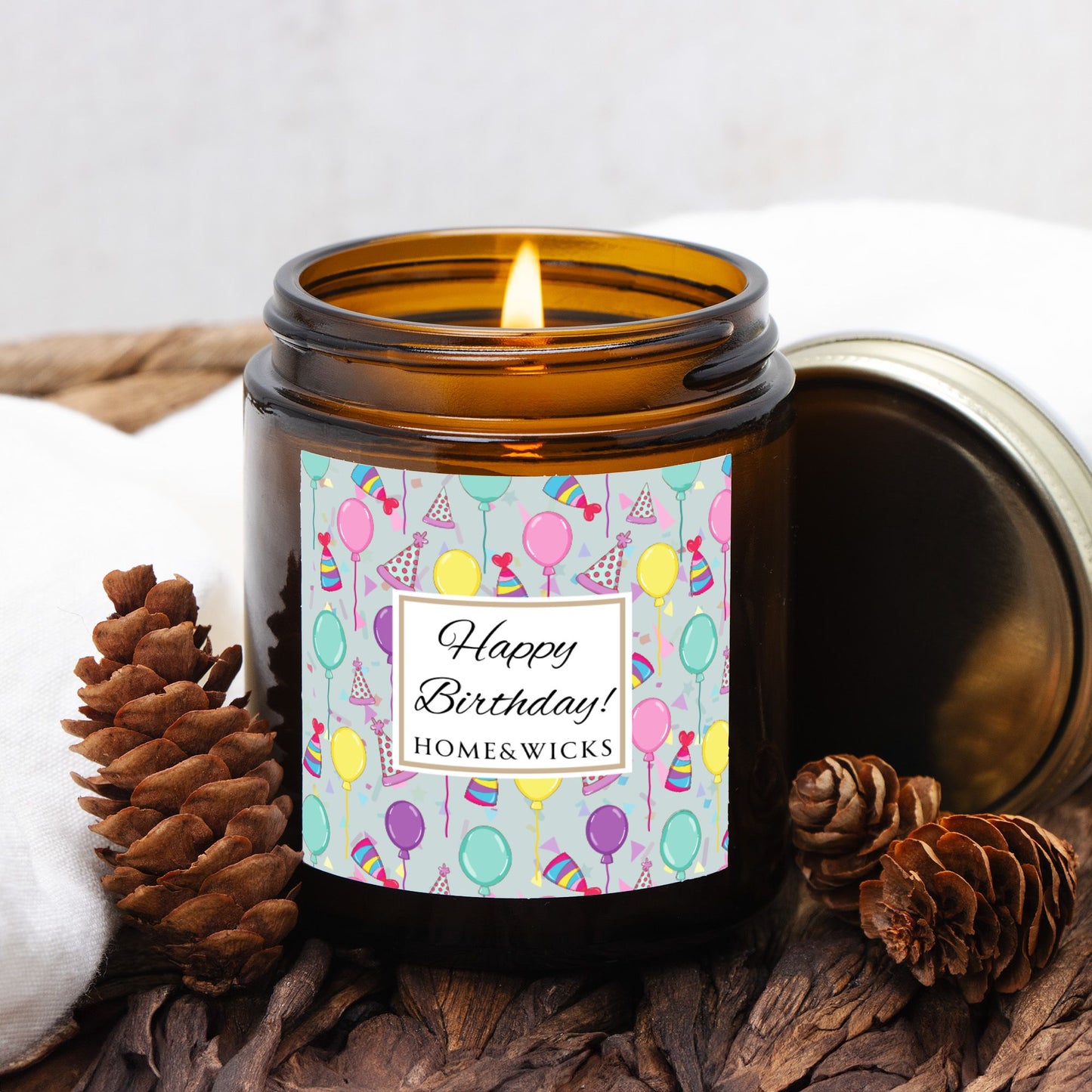 Amber candle jar with gold lid with a 'Happy Birthday' label featuring balloons, birthday party hats and confetti on a decorative background with pine cones and cotton balls.