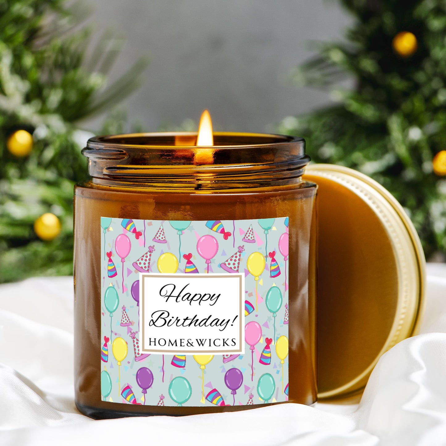 Amber candle jar with gold lid with a 'Happy Birthday' label featuring balloons, birthday party hats and confetti surrounded by greenery.
