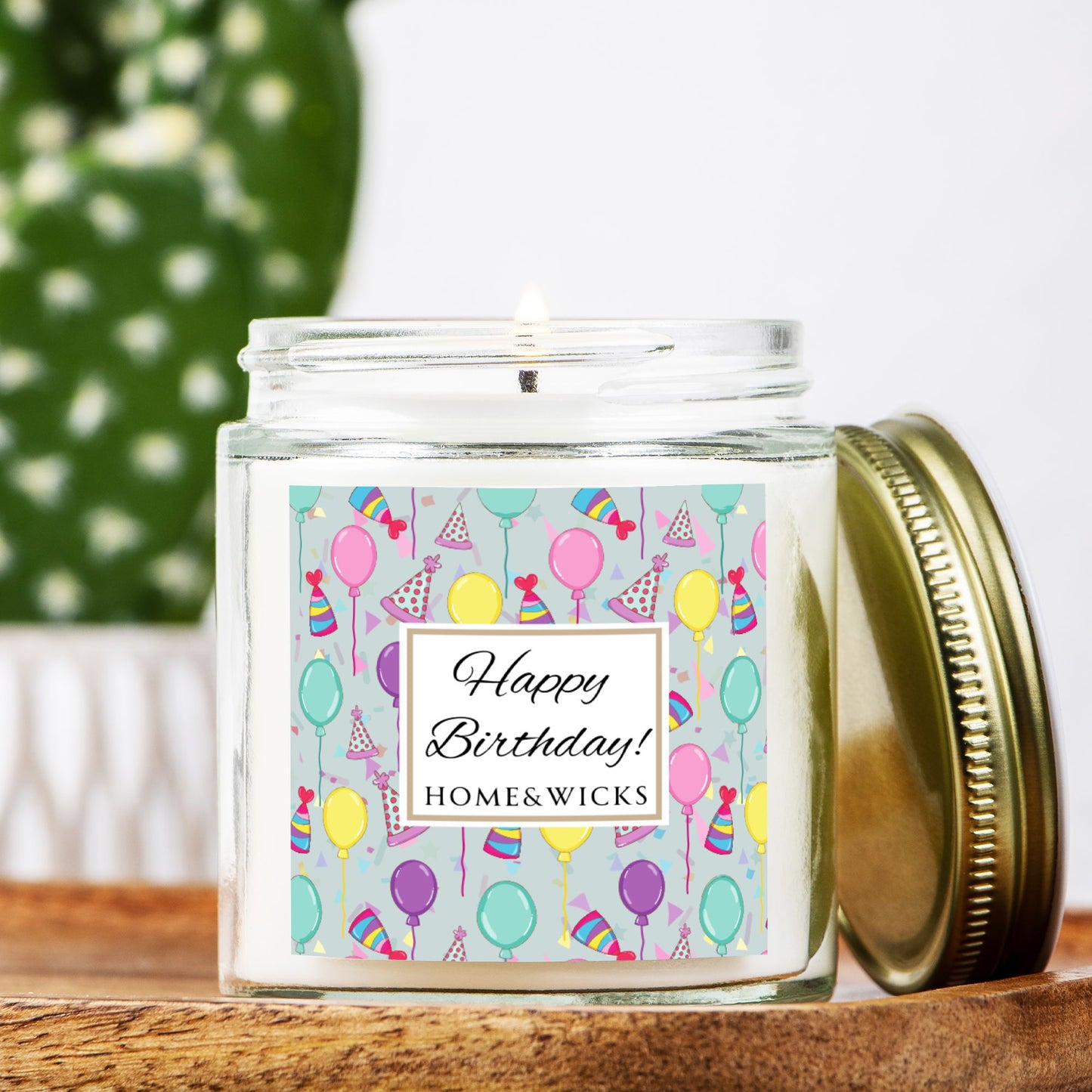 Clear candle jar with gold lid with a 'Happy Birthday' label featuring balloons, birthday party hats and confetti on a wooden surface