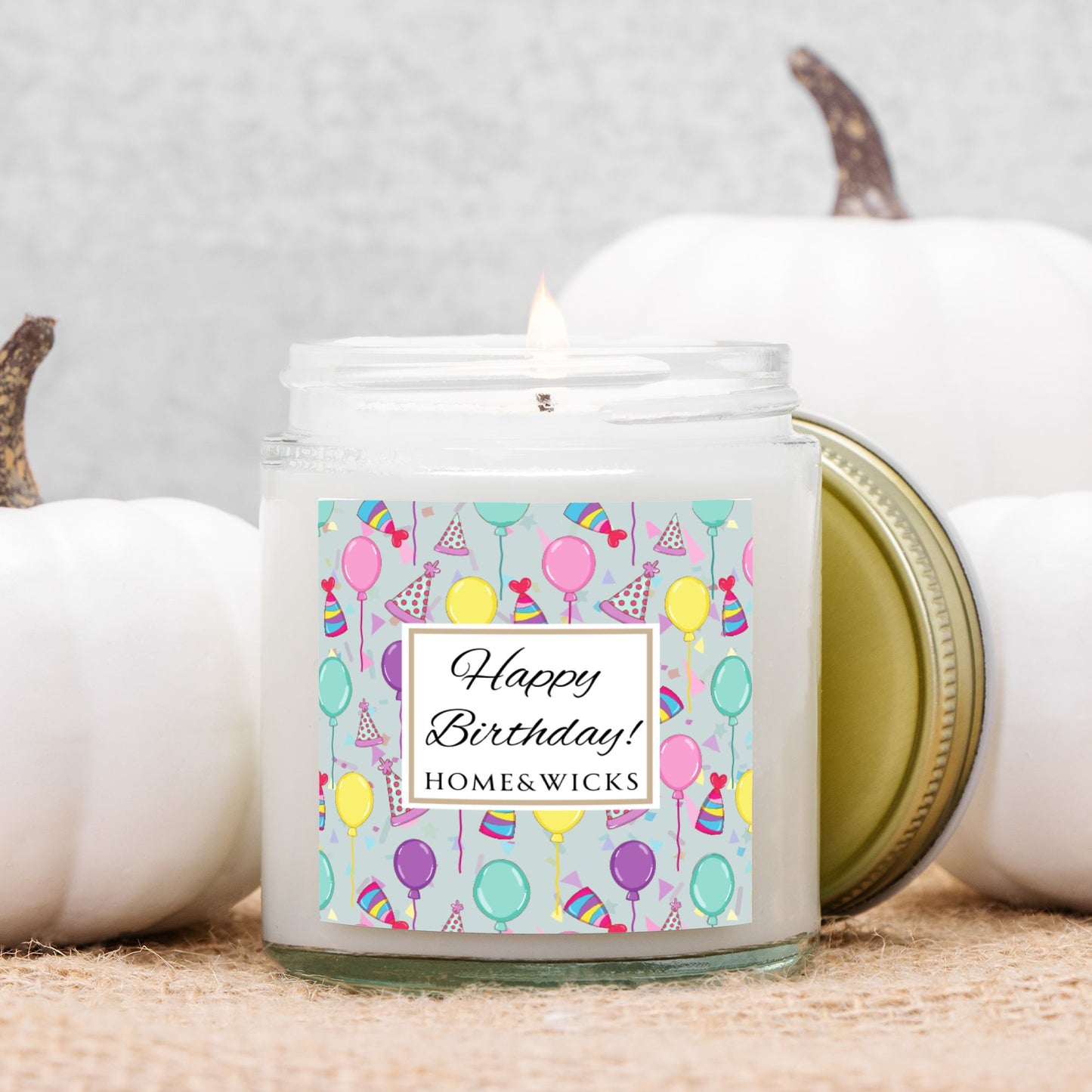 Clear candle jar with gold lid with a 'Happy Birthday' label featuring balloons, birthday party hats and confetti on a neutral background