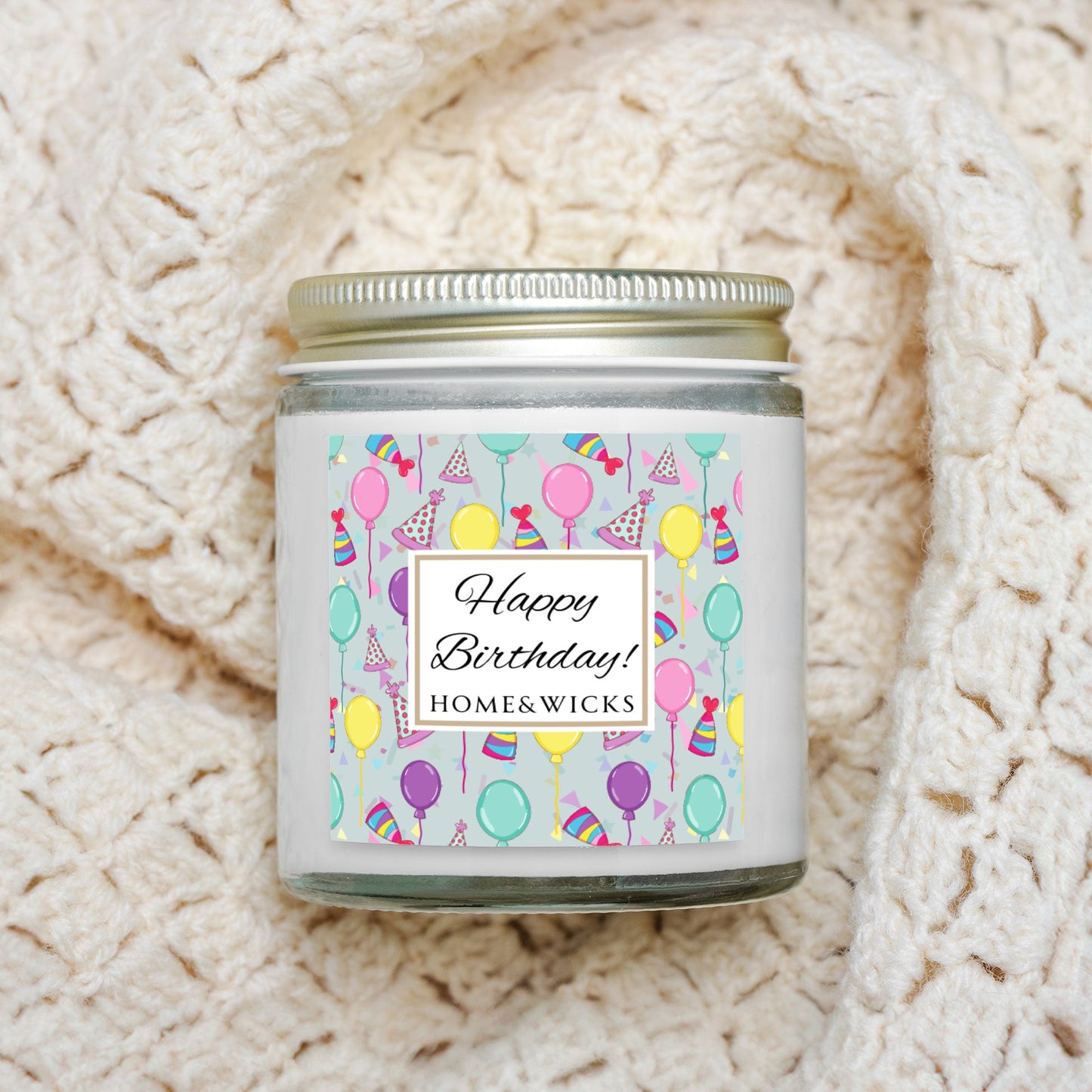 Clear candle jar with gold lid with a 'Happy Birthday' label featuring balloons, birthday party hats and confetti on a textured white background