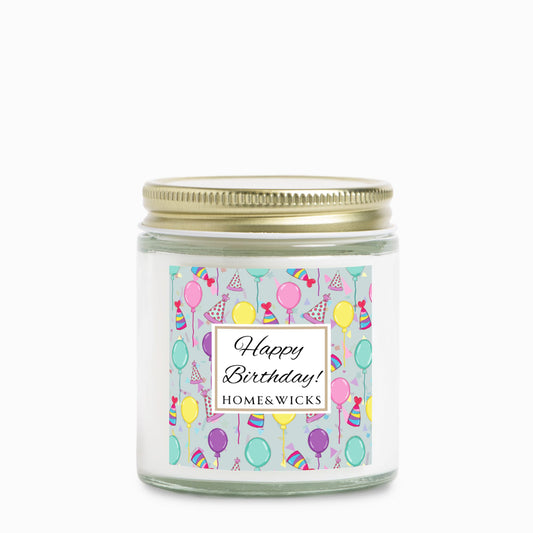 Clear candle jar with gold lid with a 'Happy Birthday' label featuring balloons, birthday party hats and confetti on white background