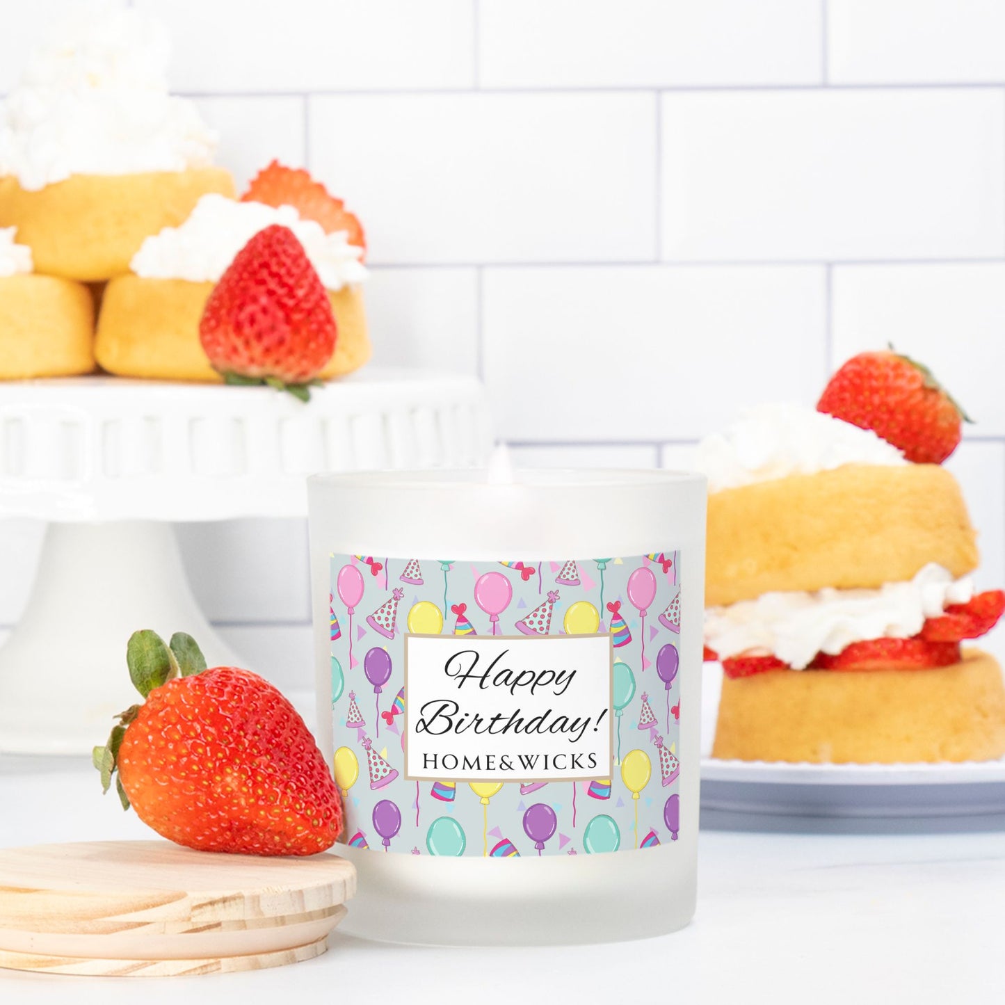 Pink wick candle in a frosted jar with bamboo wood lid and with a 'Happy Birthday' label with birthday balloons, birthday hats and confetti by Home & Wicks on a white background surrounded by strawberry shortcakes on a white tiled background