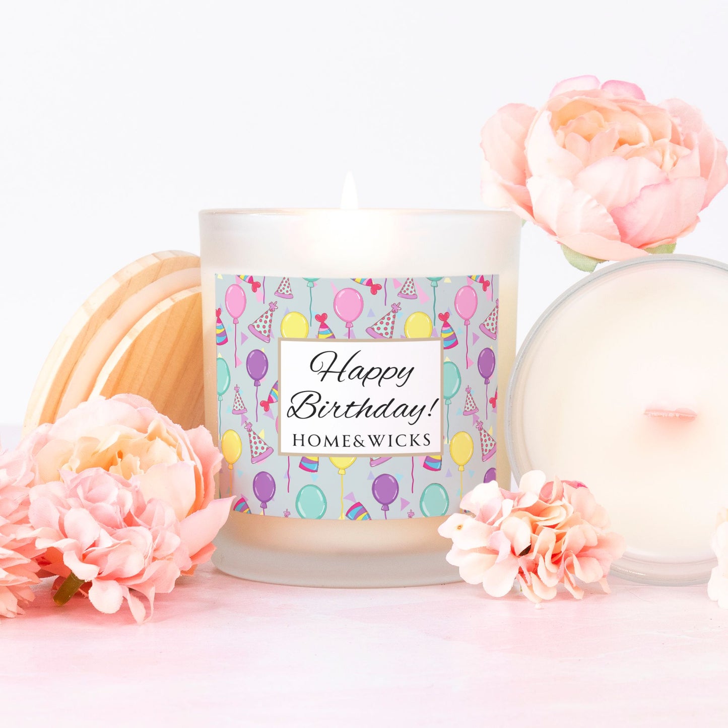 Pink wick candle in a frosted jar with bamboo wood lid and with a 'Happy Birthday' label with birthday balloons, birthday hats and confetti by Home & Wicks on a white background surrounded by flowers on a soft pink background