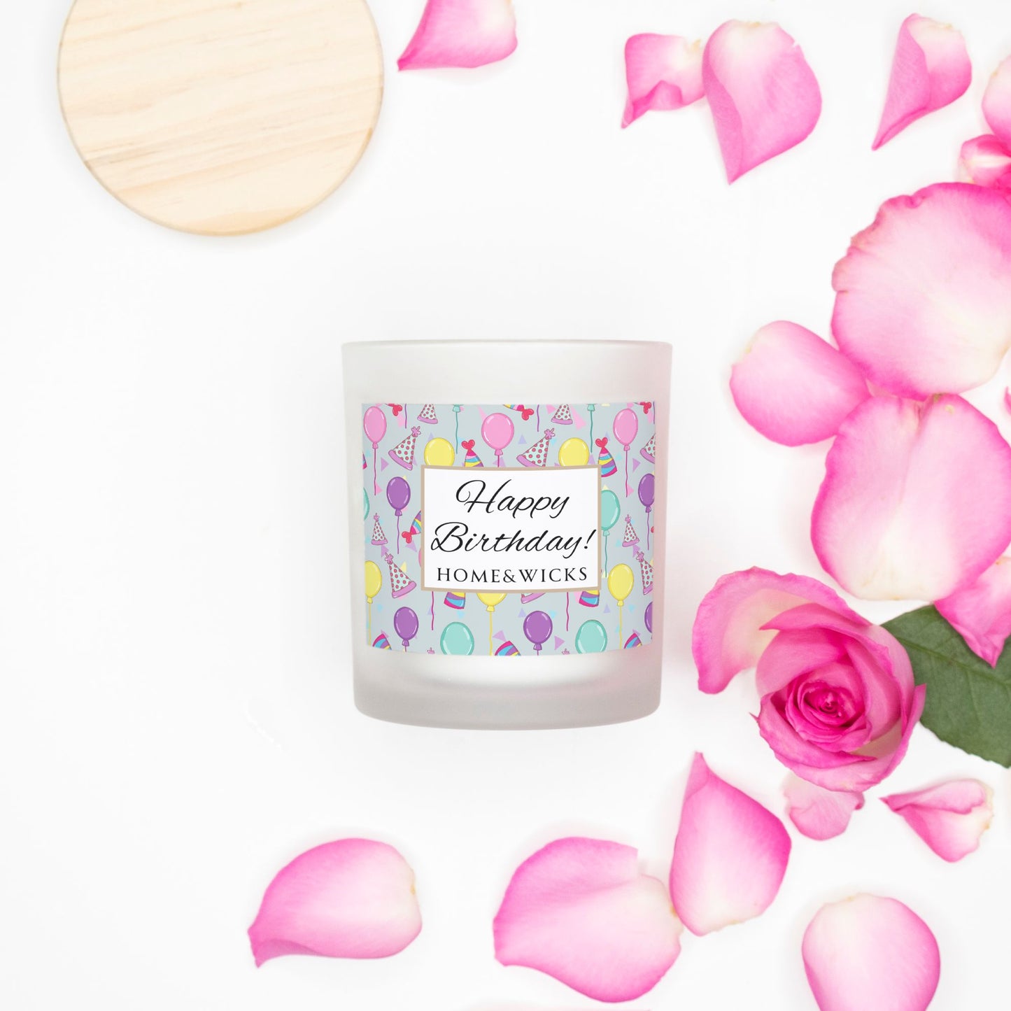 Pink wick candle in a frosted jar with bamboo wood lid and with a 'Happy Birthday' label with birthday balloons, birthday hats and confetti by Home & Wicks on a white background surrounded by pink roses and petals on a white background