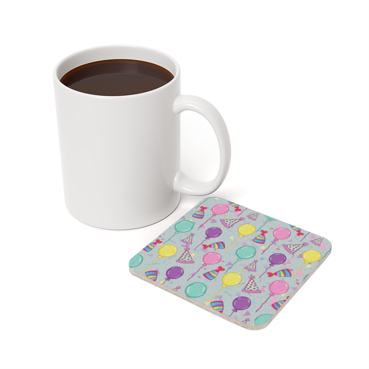 White mug with coffee on a white background with a square Happy Birthday! coaster with colorful balloons, birthday party hats and confetti pattern on a light blue gray background