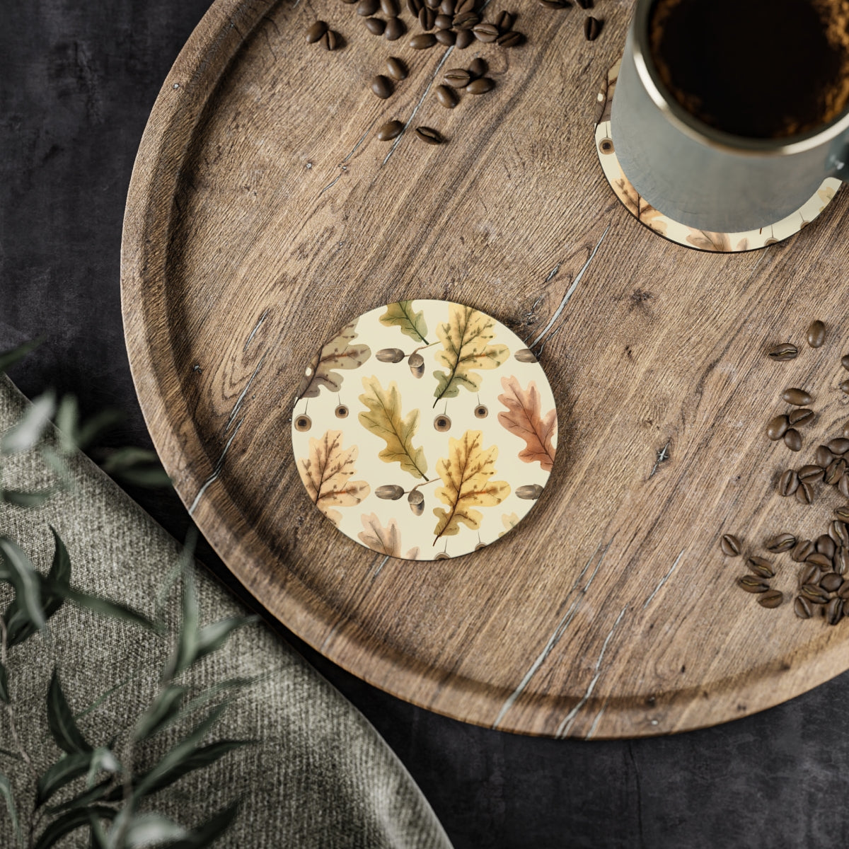 A round hardboard coaster with cork backing, featuring an oak leaf design, placed on a wooden tray with a cup of coffee and scattered coffee beans around it.