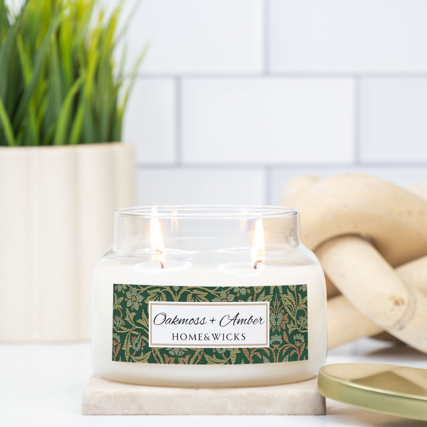 Candle in a glass jar with a Oakmoss Amber label on a white surface, surrounded by plants and wooden elements, flower garden by William Morris