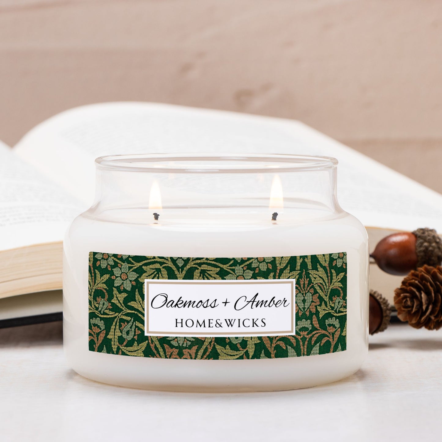 Candle in a glass jar with a decorative label with Flower Garden by William Morris on a light surface, surrounded by acorns and pine cones.