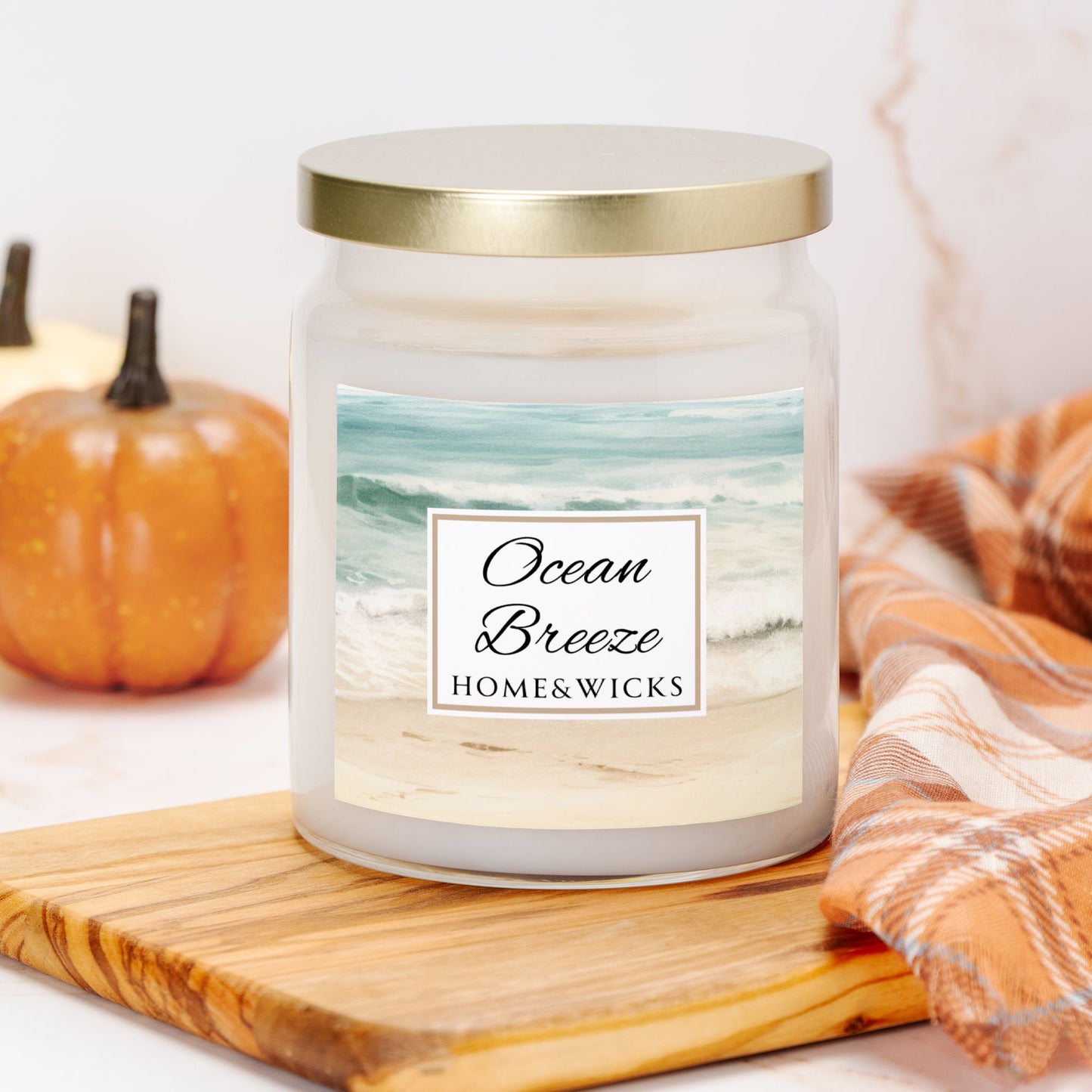 Candle labeled 'Ocean Breeze' by Home & Wicks on a wooden board with a pumpkin and plaid fabric in the background.