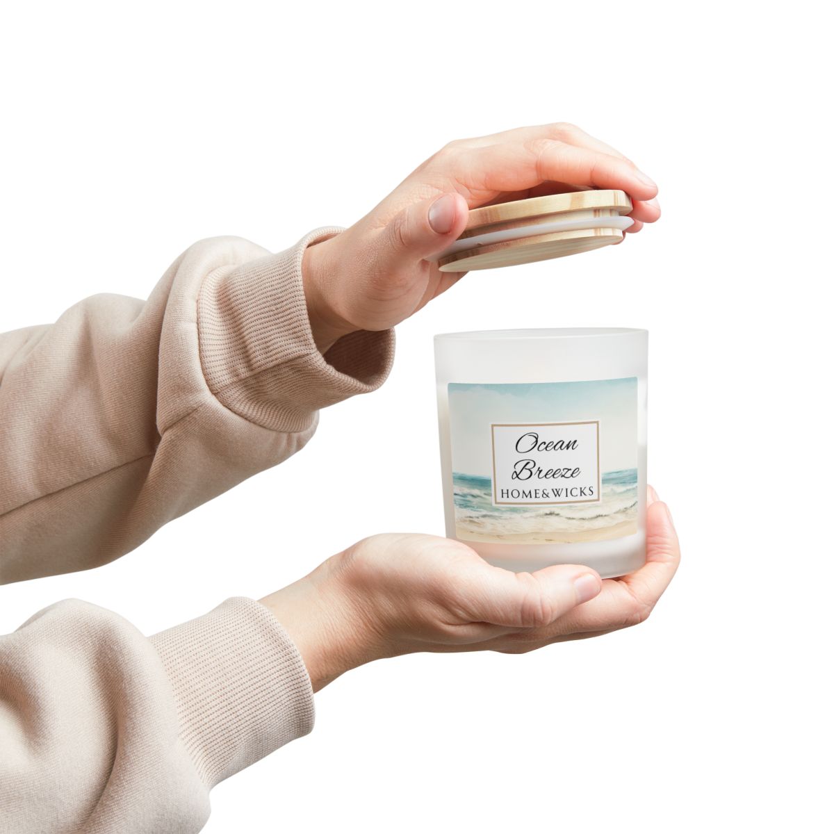 Person holding a 'Ocean Breeze' candle with ocean shore image by Home & Wicks with a beige sweater sleeve.