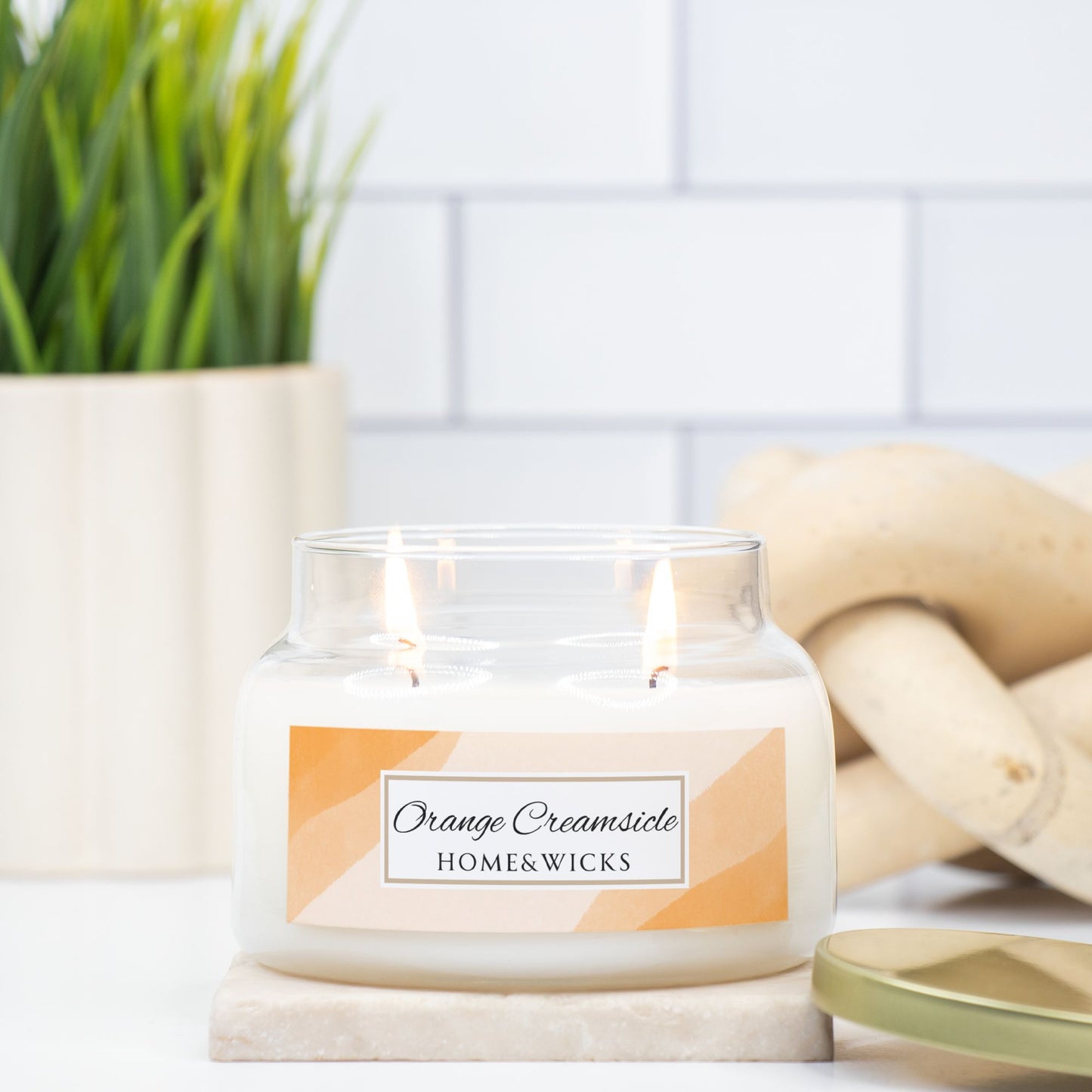 Candle labeled 'Orange Creamsicle' from HOME & WICKS on a white surface with a plant and wooden items in the background.