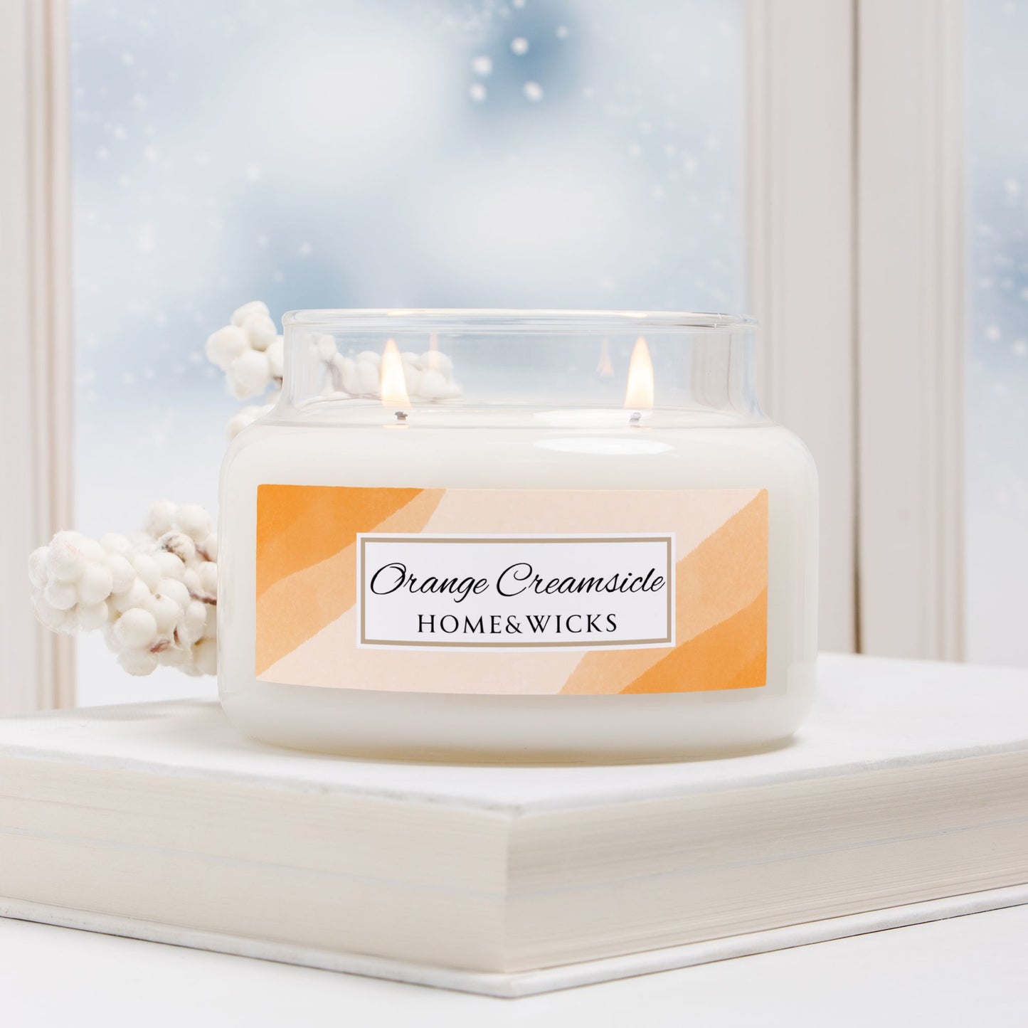 Candle labeled 'Orange Creamsicle' by HOME&WICKS on a windowsill with snow outside.