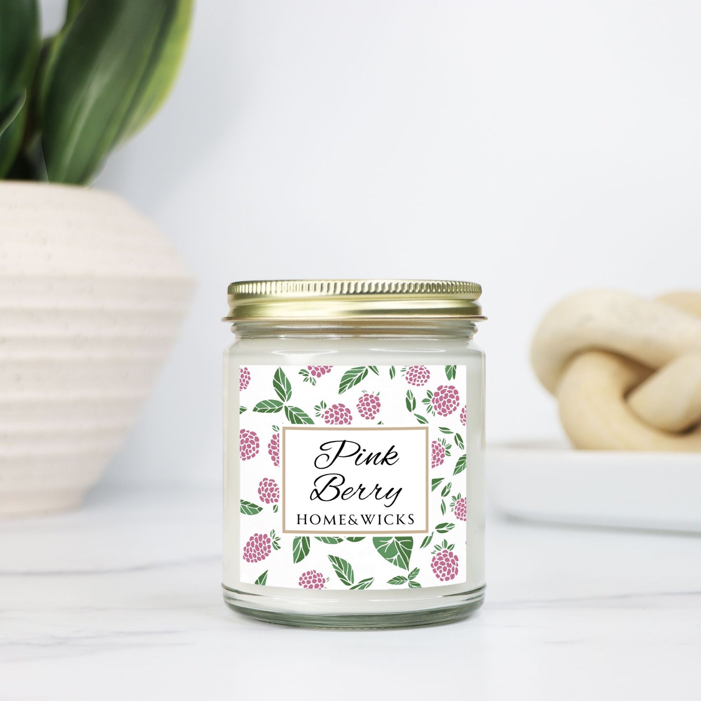 Pink Berry clear candle jar with gold lid with a decorative label featuring pink berries and green leaves on a white background by Home & Wicks on a white surface with decorative elements.