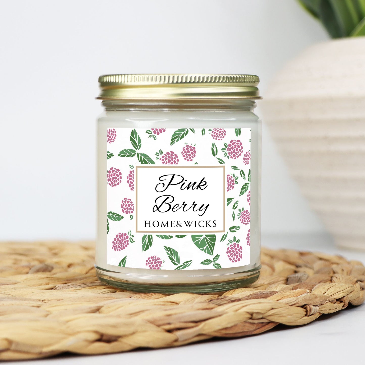 Pink Berry clear candle jar with gold lid with a decorative label featuring pink berries and green leaves on a white background by Home & Wicks on a woven mat with a white background