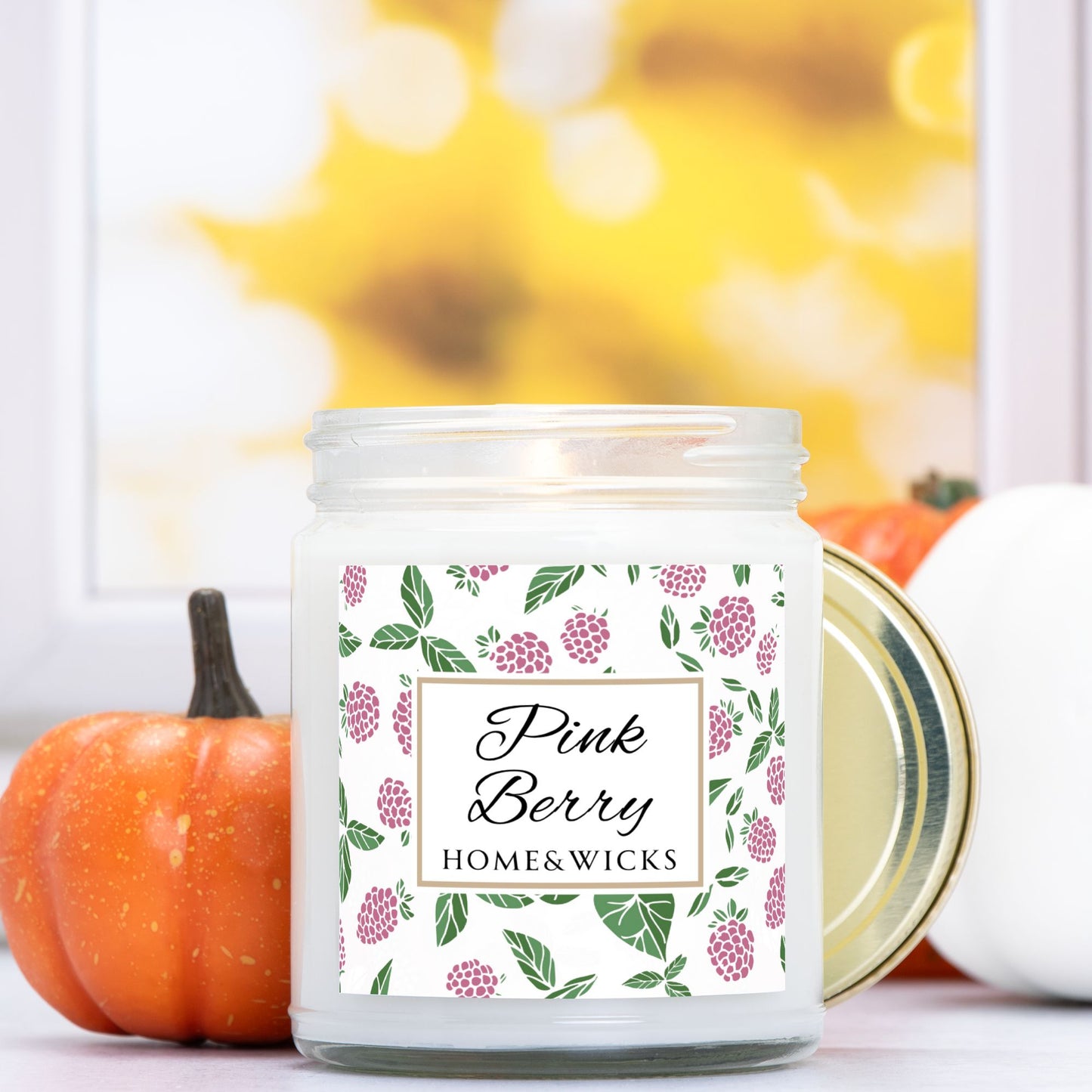 Pink Berry clear candle jar with gold lid with a decorative label featuring pink berries and green leaves on a white background by Home & Wicks, set against a blurred background with pumpkins.