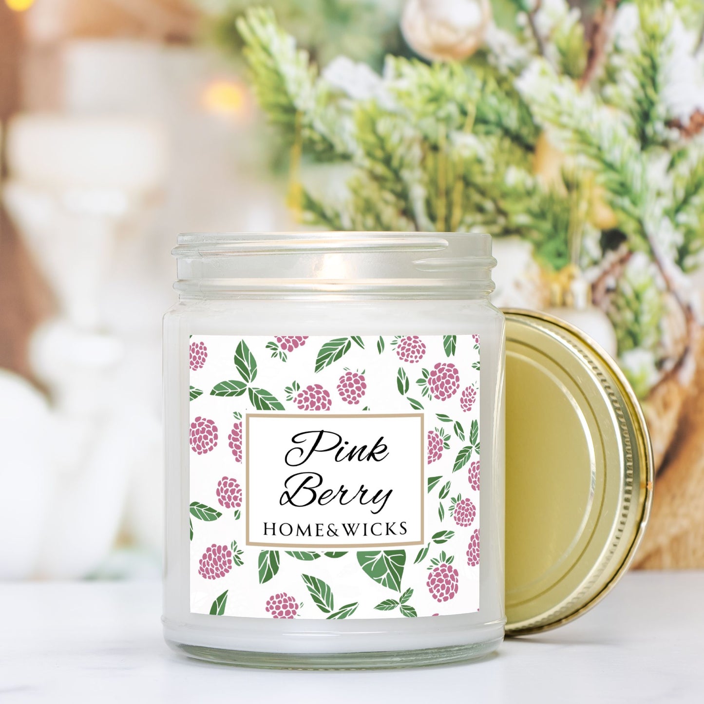 Pink Berry clear candle jar with gold lid with a decorative label featuring pink berries and green leaves on a white background by Home & Wicks on a blurred background with greenery