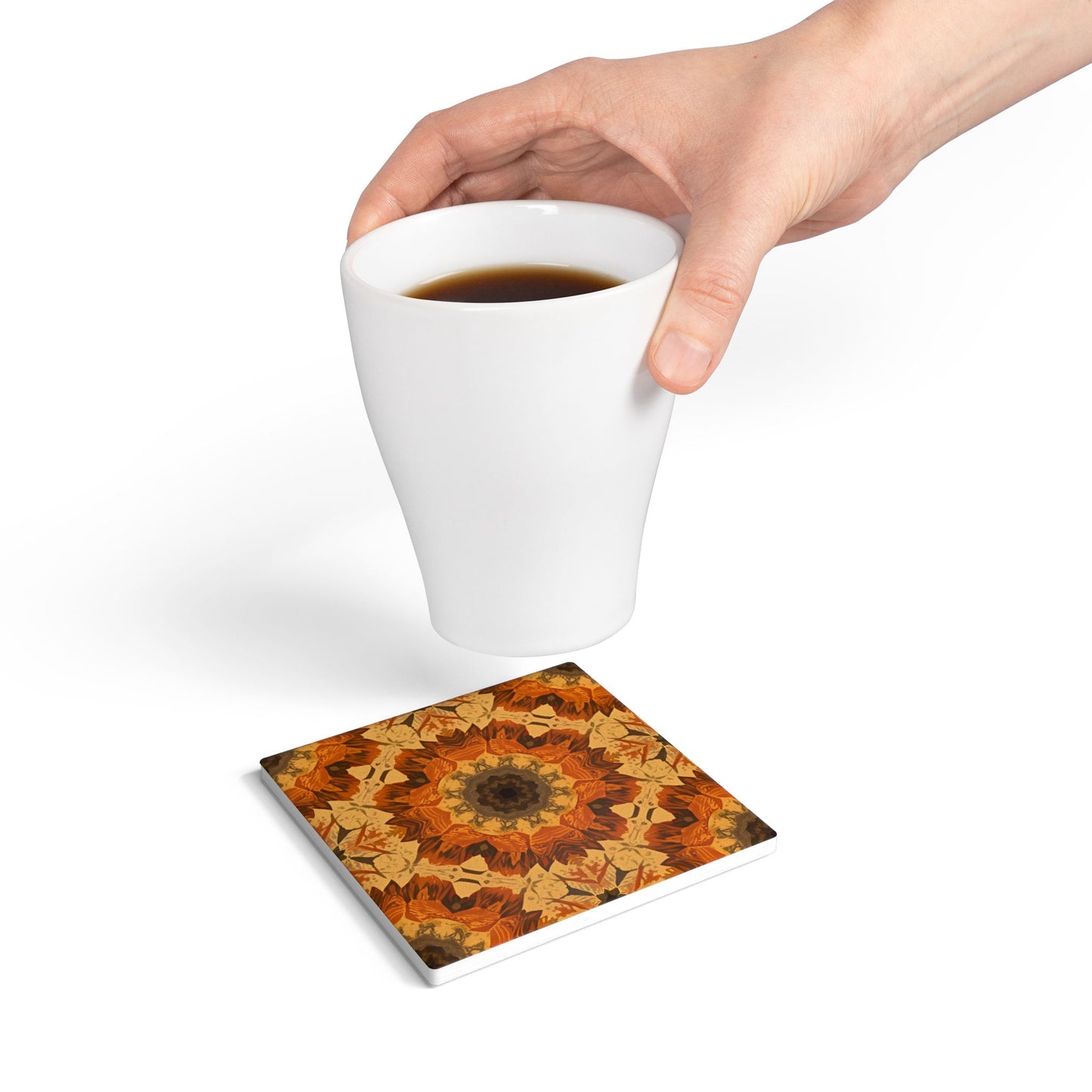 Hand holding a white mug with a mandala orange, yellow and gray tile coaster underneath on a white background