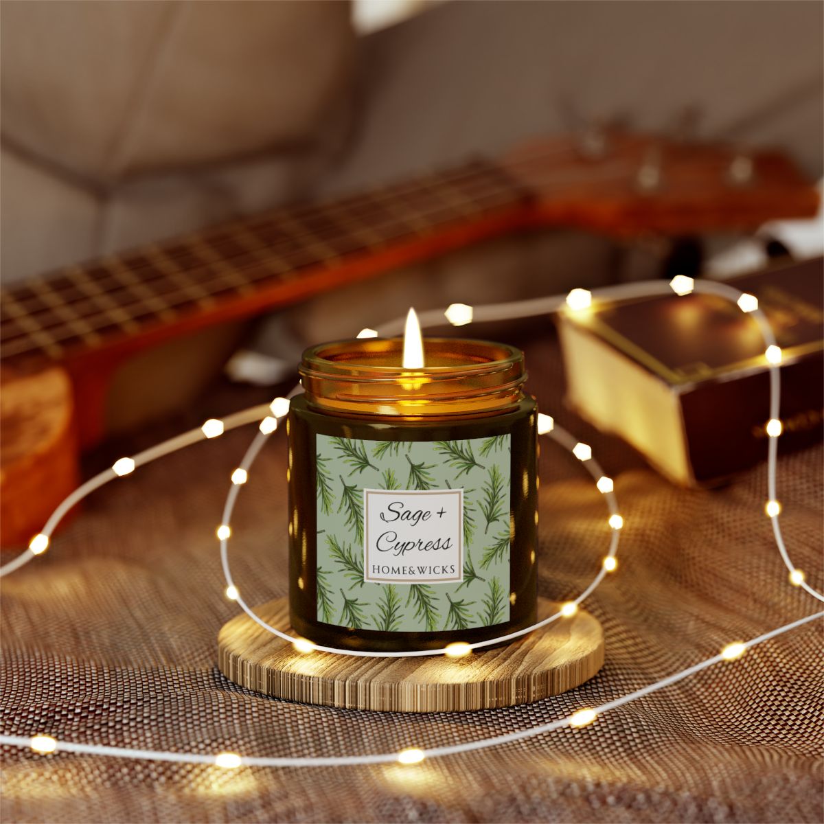 Sage & Cypress candle in a 4 oz. amber glass jar with a gold lid and 'Sage + Cypress' label on a white background by Home & Wicks on a wooden coaster with string lights and a guitar in the background.