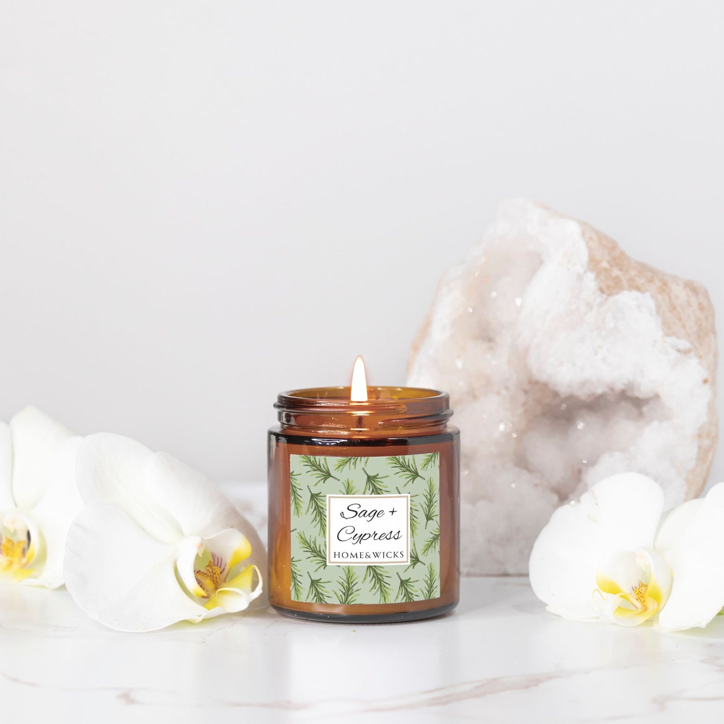 Sage & Cypress candle in a 4 oz. amber glass jar with a gold lid and 'Sage + Cypress' label on a white background by Home & Wicks in front of white flowers and a crystal on a light background