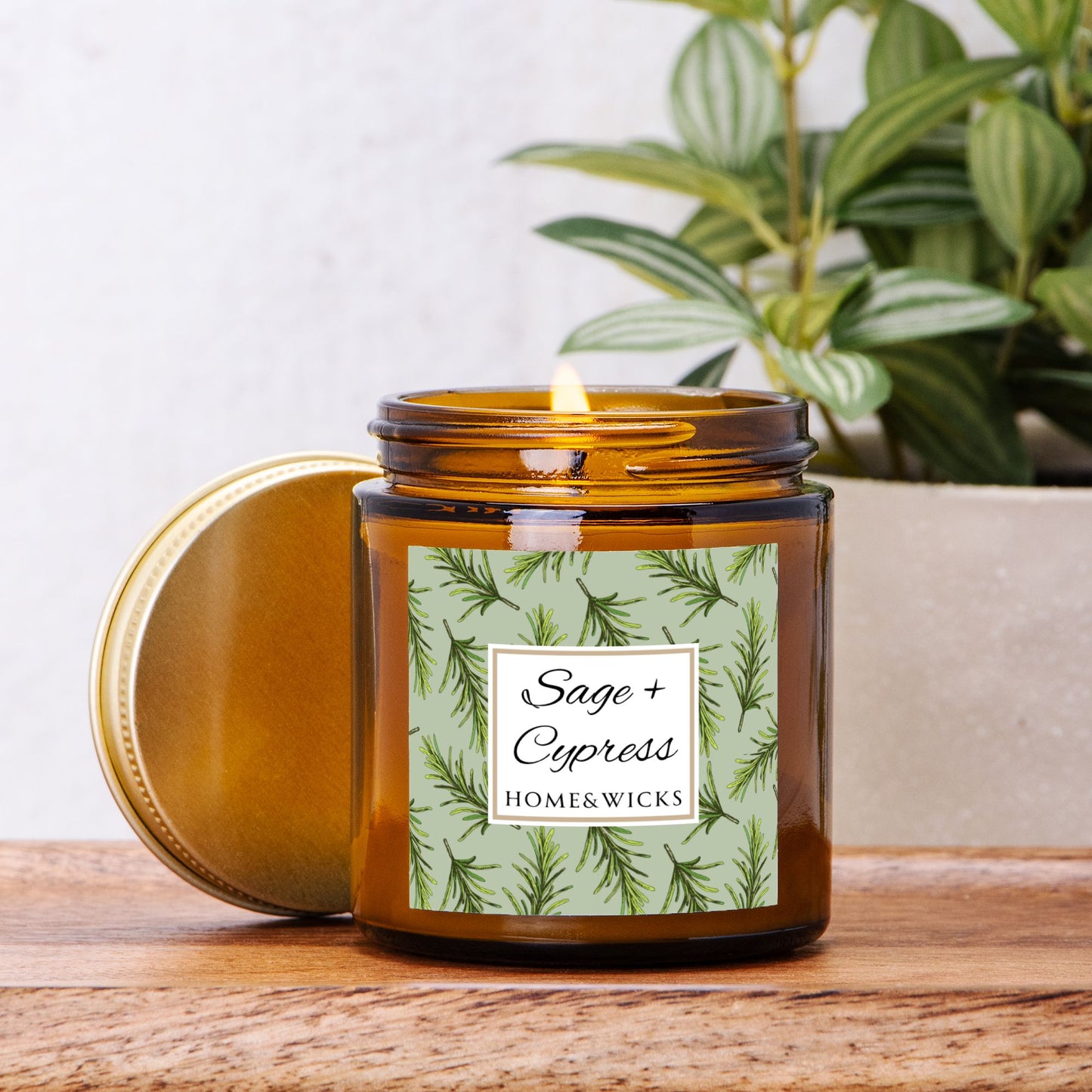 Sage & Cypress candle in a 4 oz. amber glass jar with a gold lid and 'Sage + Cypress' label on a white background by Home & Wicks on a wooden surface with a plant in the background.