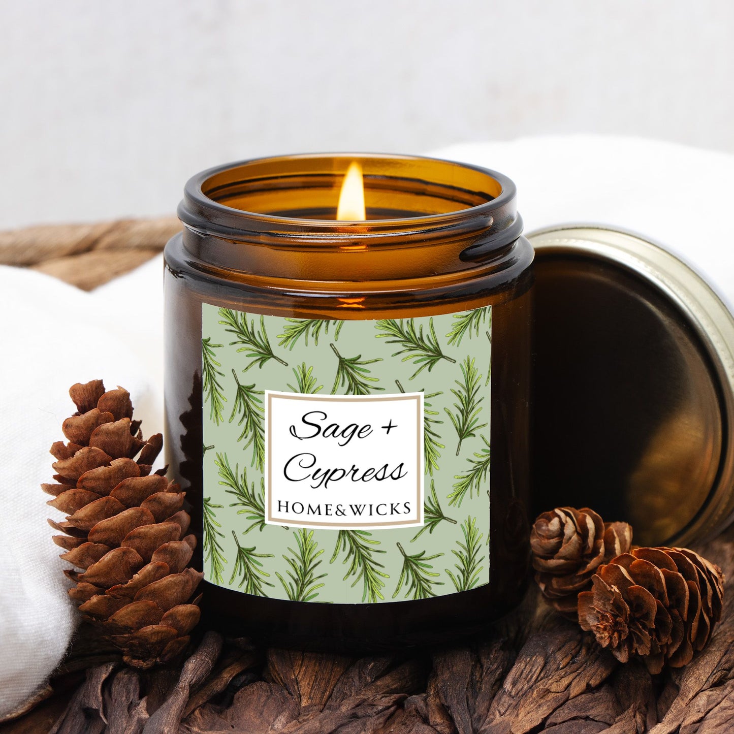 Sage & Cypress candle in a 4 oz. amber glass jar with a gold lid and 'Sage + Cypress' label on a white background by Home & Wicks surrounded by pine cones and wood.