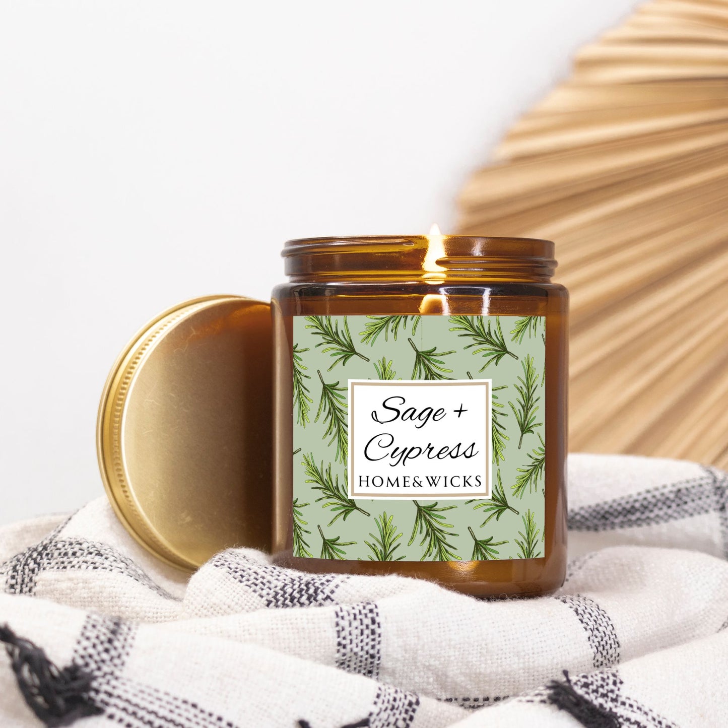 Sage & Cypress candle in an amber jar with gold lid with 'Sage + Cypress' label on a light green background with cypress needle leaves on a white and black checkered cloth.
