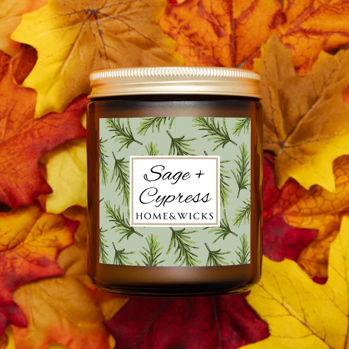 Sage & Cypress candle in an amber jar with gold lid with 'Sage + Cypress' label on a light green background with cypress needle leaves on a bed of autumn leaves.