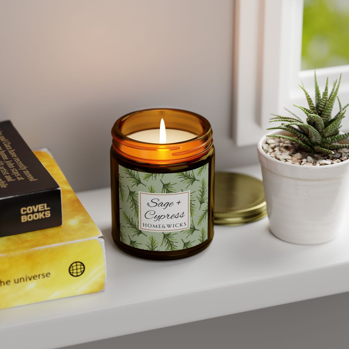Sage & Cypress candle in an amber jar with gold lid with 'Sage + Cypress' label on a light green background with cypress needle leaves on a shelf next to books and a plant
