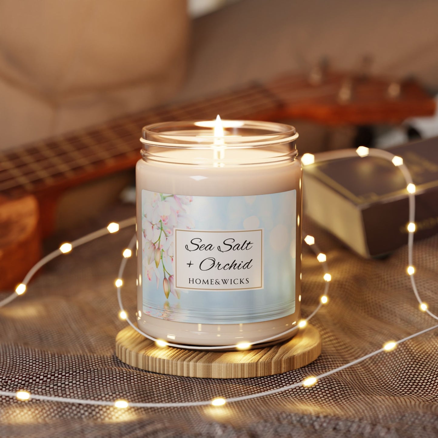 Candle labeled 'Sea Salt + Orchid' by Homewicks on a wooden coaster with string lights and a guitar in the background.