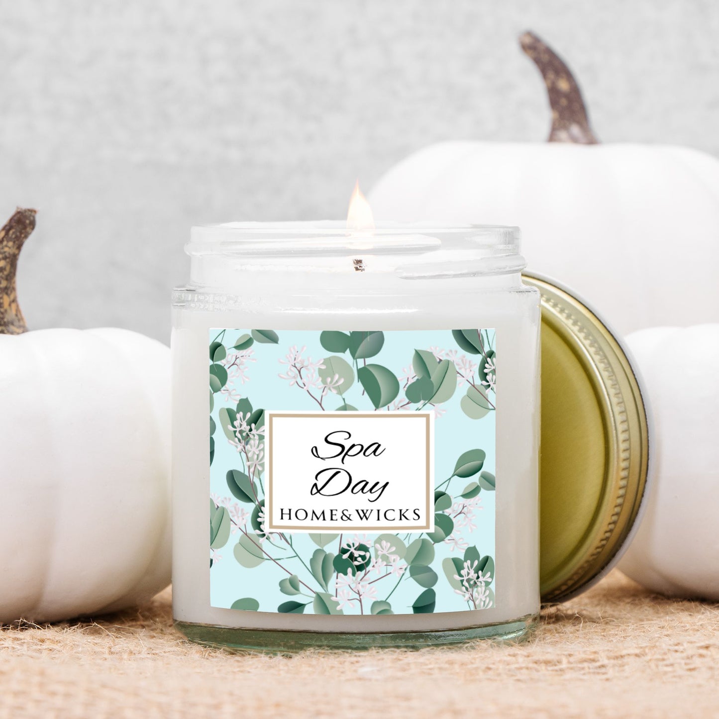 Spa Day candle by Homes & Wicks in a clear candle jar with gold lid with a eucalyptus floral label on a white background in front of white pumpkins