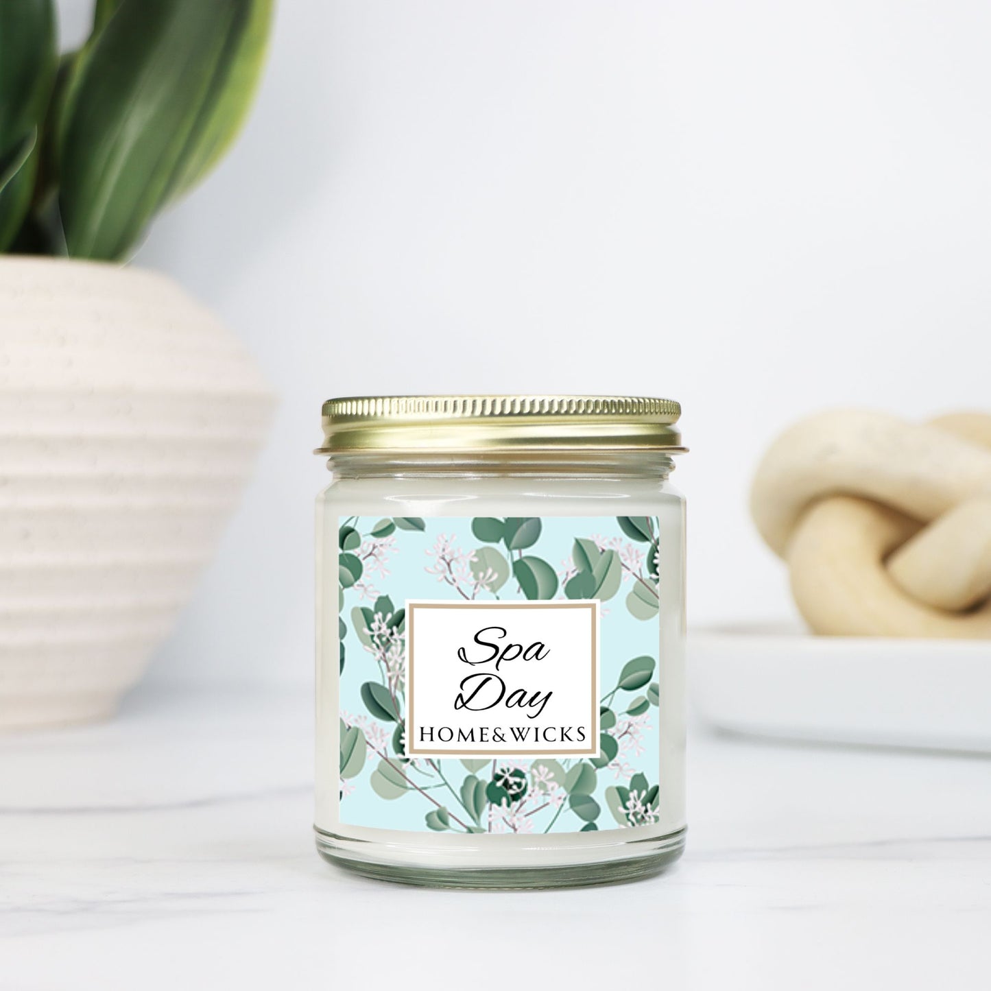 Spa Day candle clear jar with gold lid with a decorative eucalyptus label on a white background by Home & Wicks on a white surface with a plant and towel in the background.