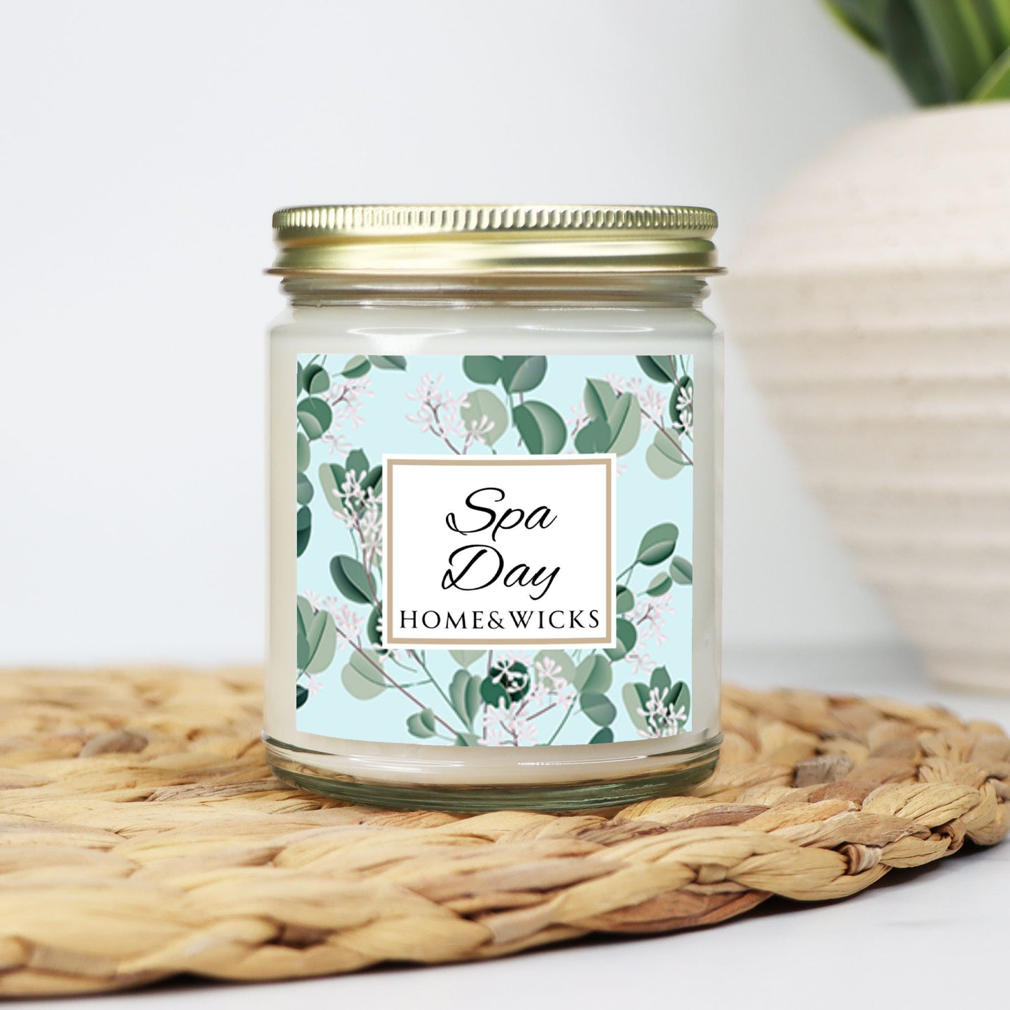 Spa Day candle clear jar with gold lid with a decorative eucalyptus label on a white background by Home & Wicks on a woven mat