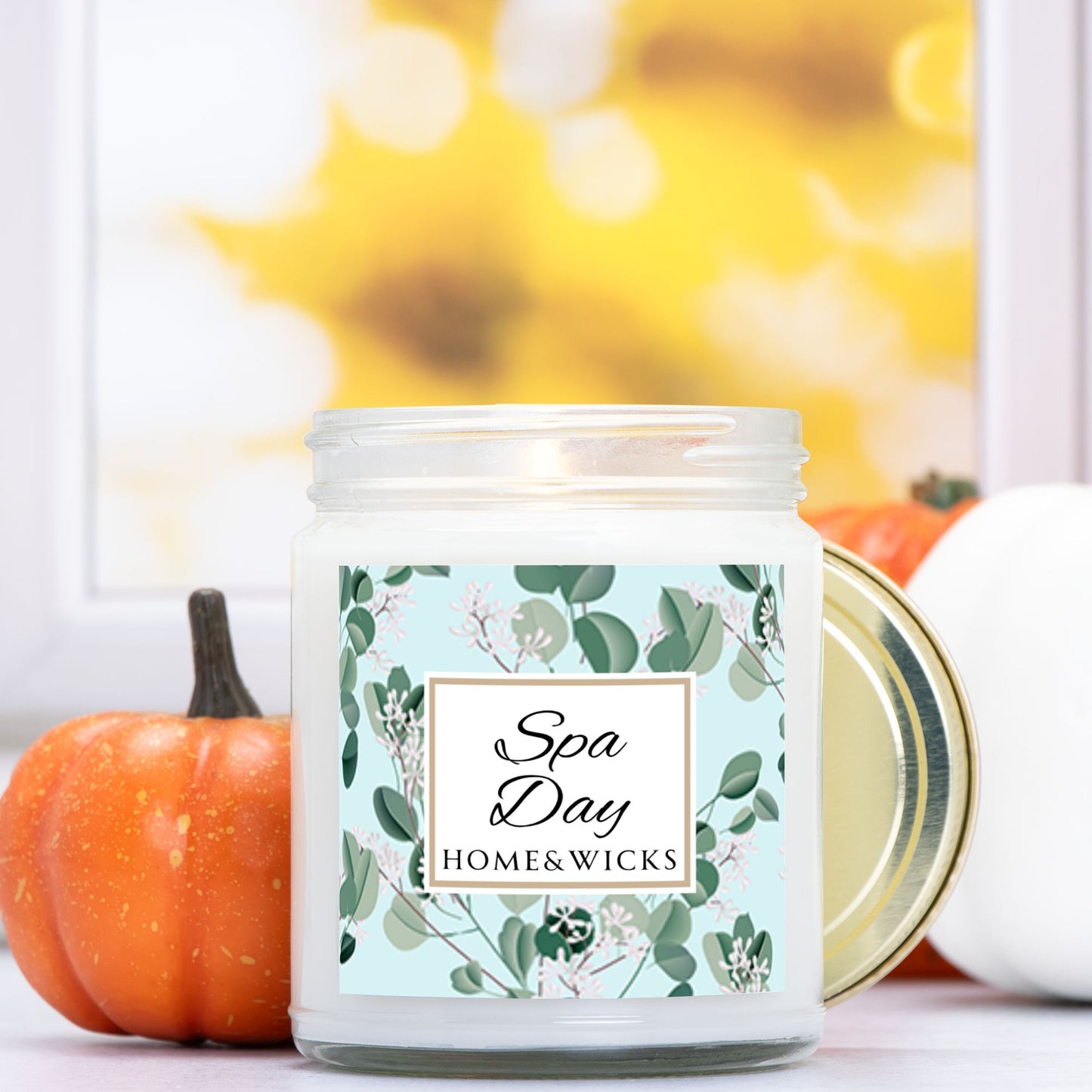 Spa Day candle clear jar with gold lid with a decorative eucalyptus label on a white background by Home & Wicks, surrounded by pumpkins and a blurred background.