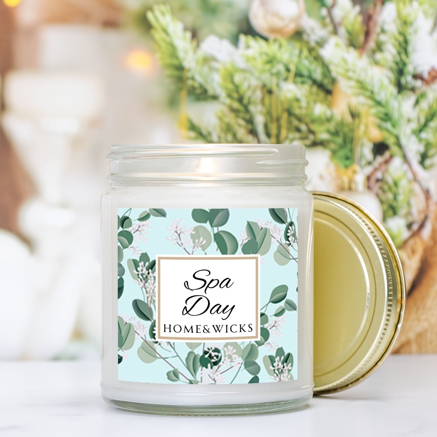 Spa Day candle clear jar with gold lid with a decorative eucalyptus label on a white background by Home & Wicks on a blurred background with greenery