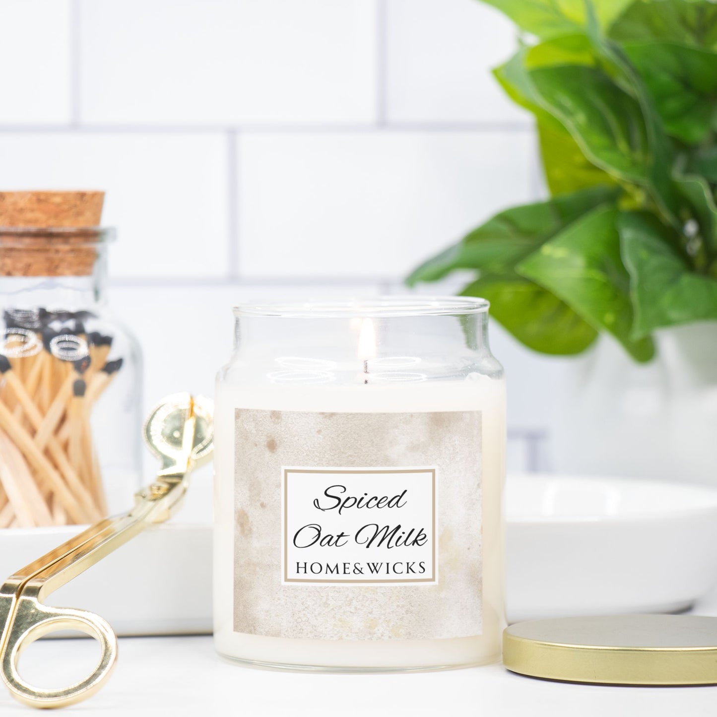 Candle jar labeled 'Spiced Oat Milk' by HOME&WICKS on a white surface with a plant and matches in the background.