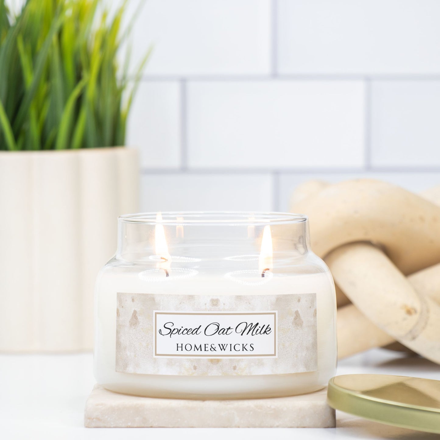 Candle labeled 'Spiced Oat Milk' from HOME & WICKS on a marble surface with a plant and wooden elements in the background.