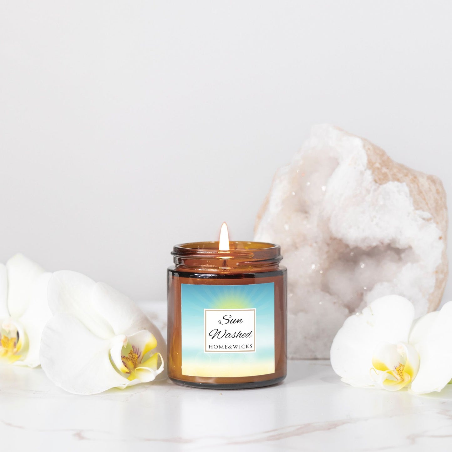 Sun Washed candle in a 4 oz. jar labeled 'Sun Washed' with sun graphic by Home & Wicks on a white background, surrounded by white flowers and a crystal on a light background