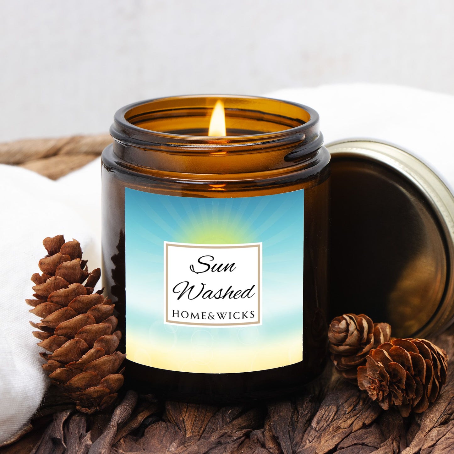 Sun Washed candle in a 4 oz. jar labeled 'Sun Washed' with sun graphic by Home & Wicks on a white background, surrounded by pine cones and wood.