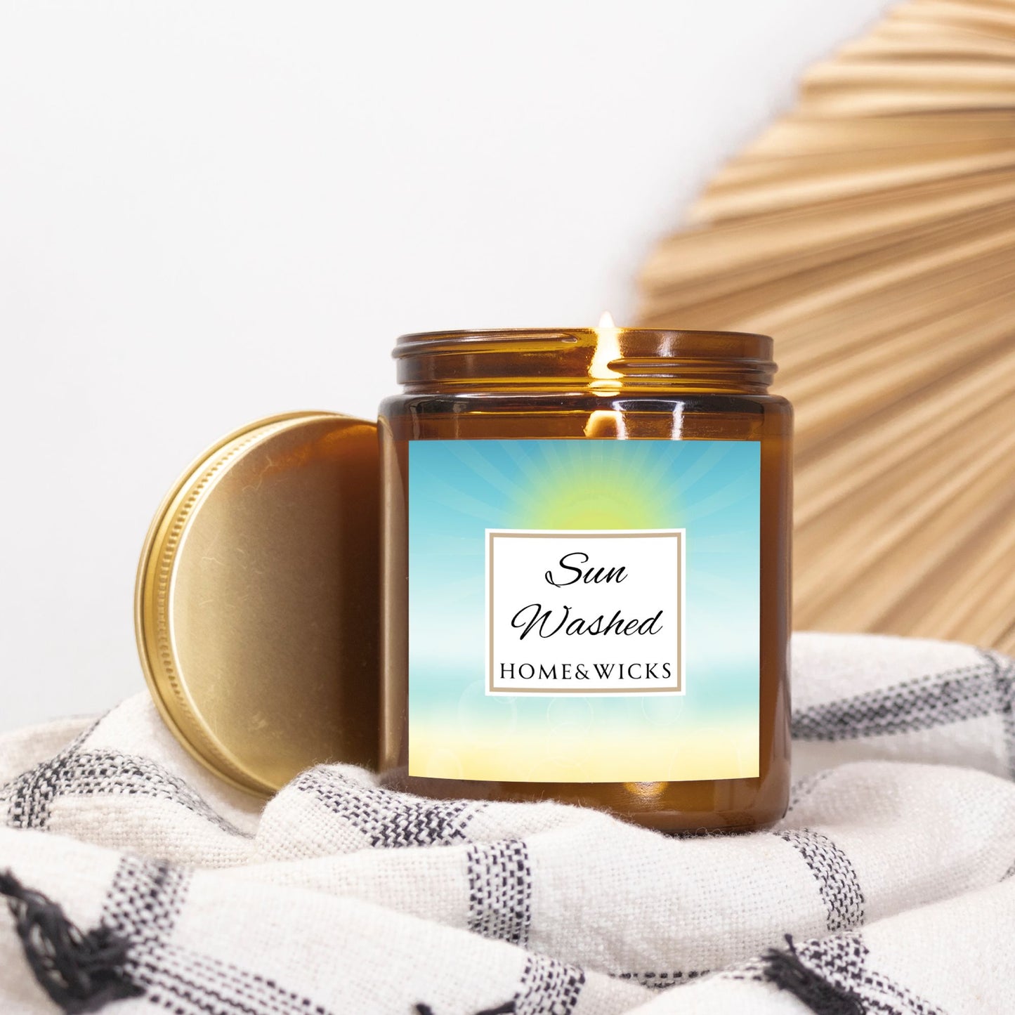 Candle in a 9 oz. amber jar with gold lid with 'Sun Washed' label on with sun graphic gradient background by Home & Wicks on a white and black patterned fabric background