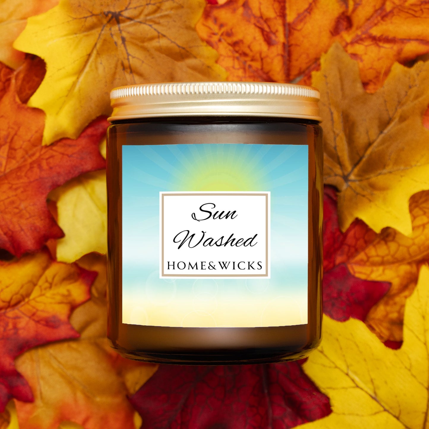 Candle in a 9 oz. amber jar with gold lid with 'Sun Washed' label on with sun graphic gradient background by Home & Wicks on a bed of autumn leaves.