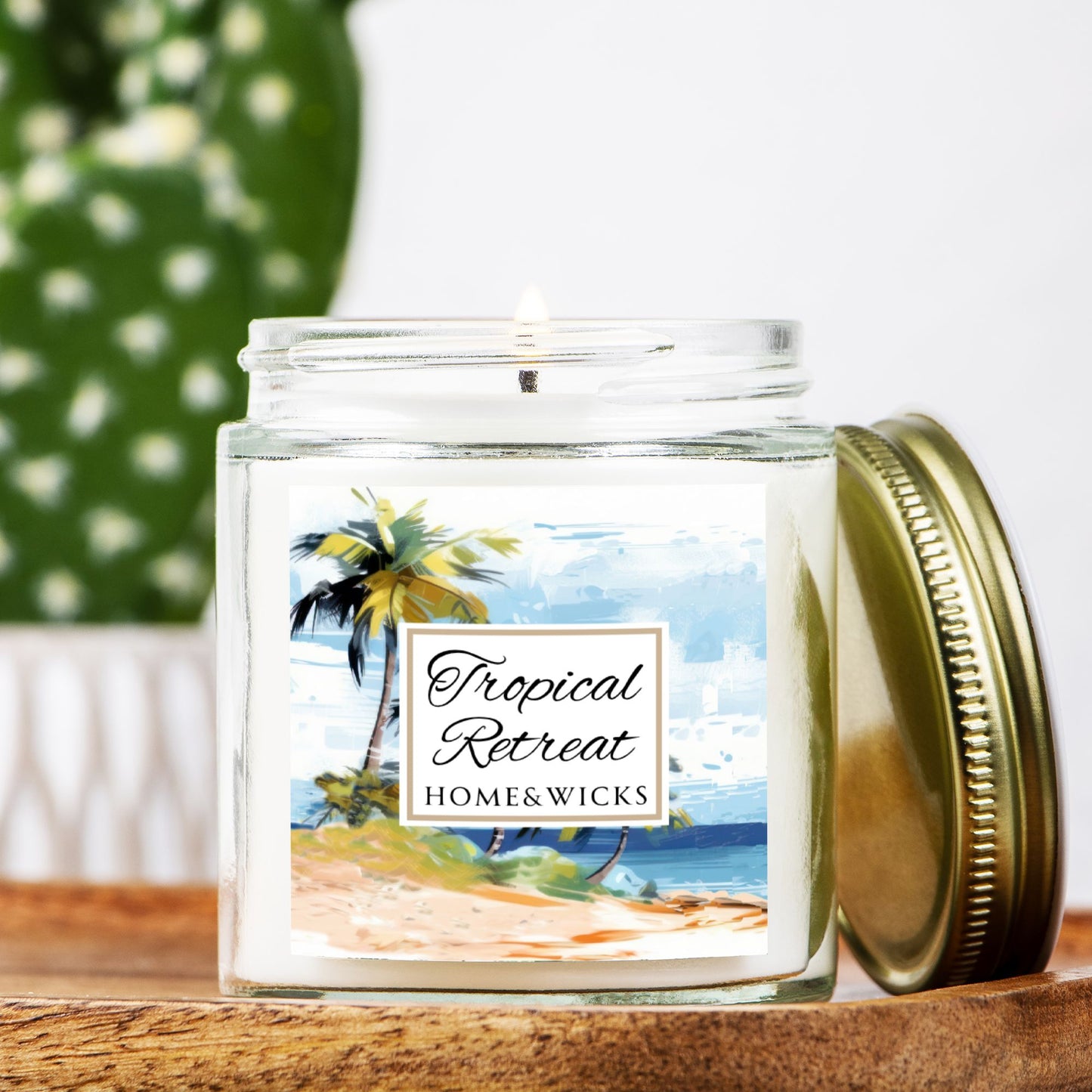 Homes & Wicks candle labeled 'Tropical Retreat' with a beach scene on a white background in a clear jar with gold lid on a wooden surface with a blurred green plant in the background.