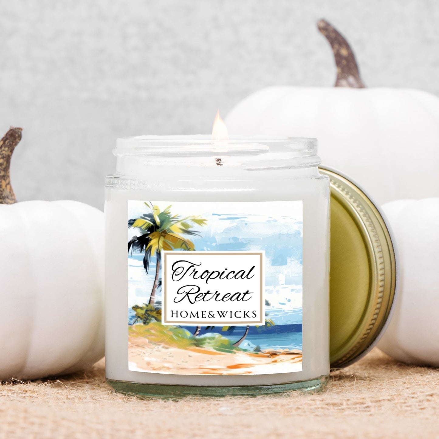 Homes & Wicks candle labeled 'Tropical Retreat' with a beach scene on a white background in a clear jar with gold lid with pumpkins in the background