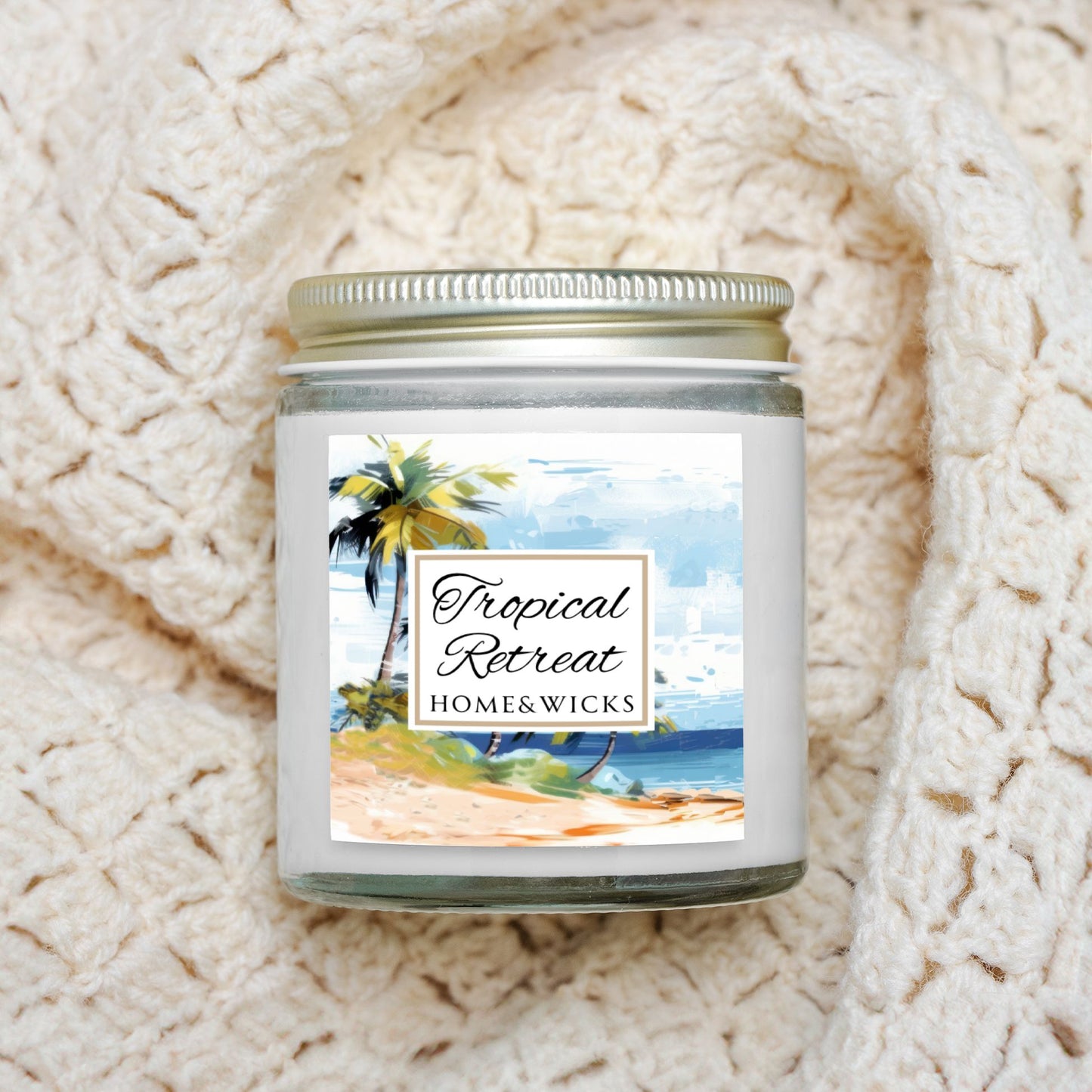 Homes & Wicks candle labeled 'Tropical Retreat' with a beach scene in a clear jar with gold lid on a textured white blanket background