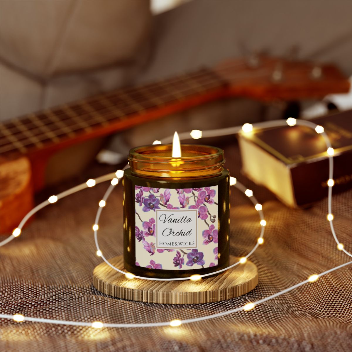 Vanilla Orchid candle in a 4 oz. amber glass candle jar with gold lid with a pink and purple orchid floral label on a white background by Home & Wicks on a wooden coaster surrounded by string lights, with a guitar in the background.