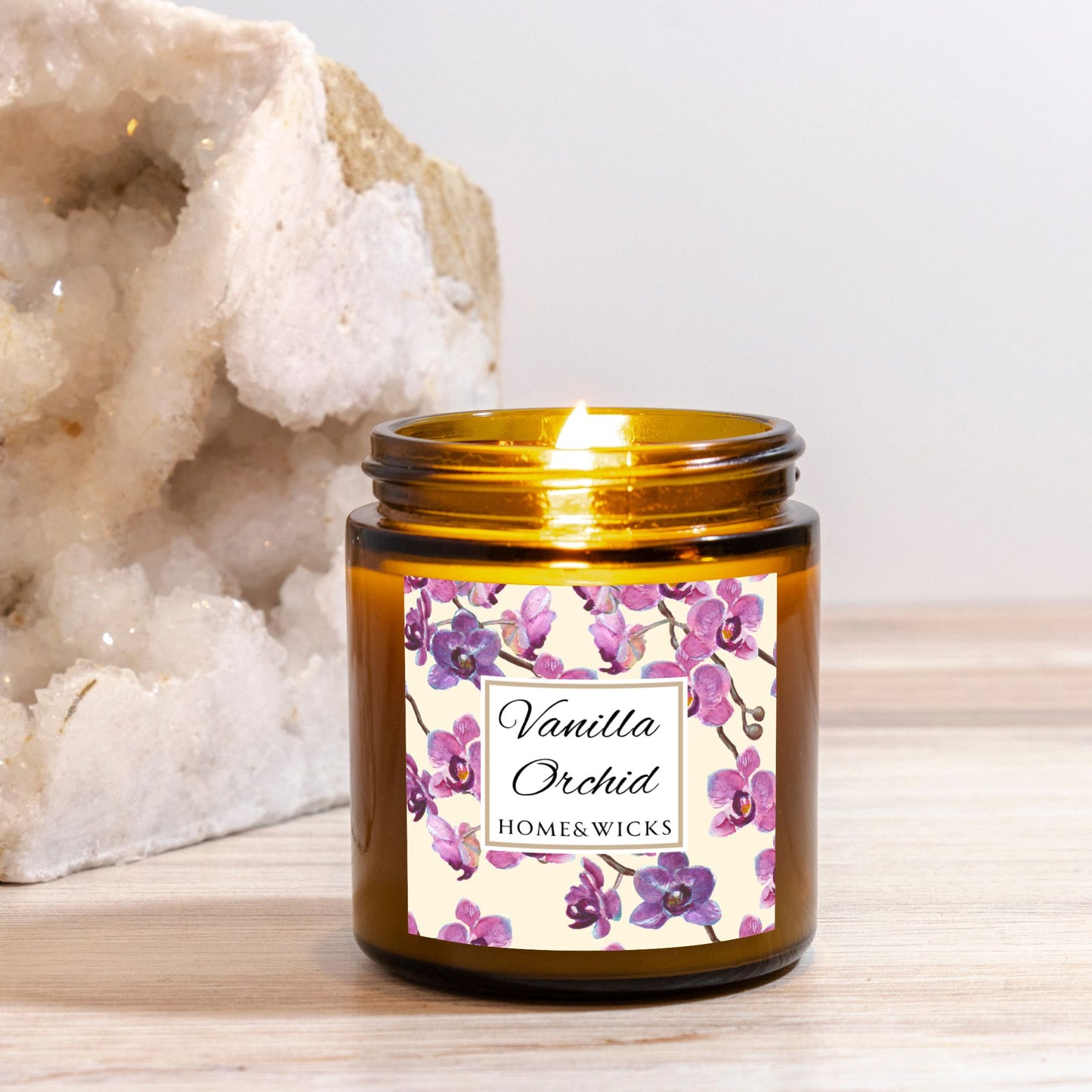 Lit Vanilla Orchid candle in a 4 oz. amber glass candle jar with gold lid with a pink and purple orchid floral label on a white background by Home & Wicks on a wooden surface.