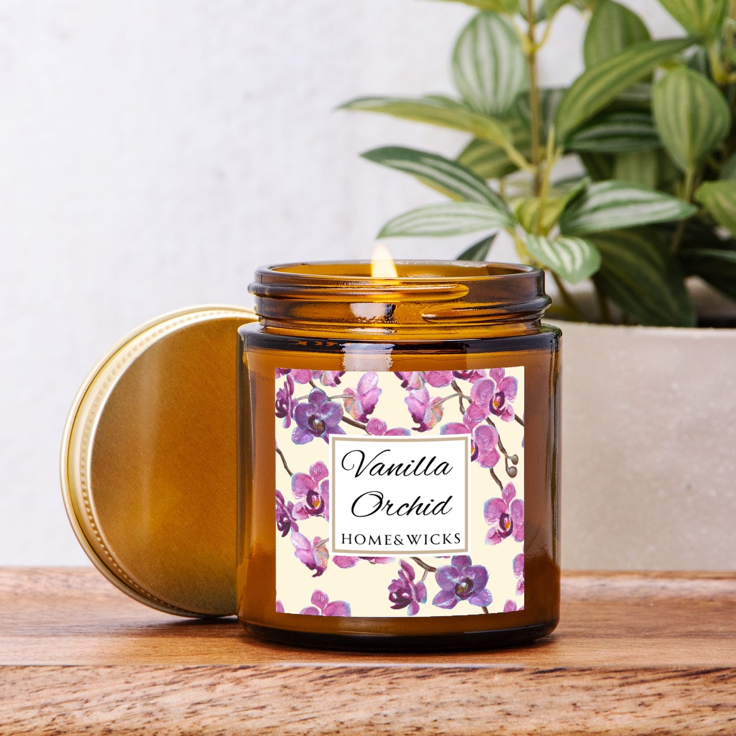 Vanilla Orchid candle in a 4 oz. amber glass candle jar with gold lid with a pink and purple orchid floral label on a white background by Home & Wicks on a wooden surface, plant in background